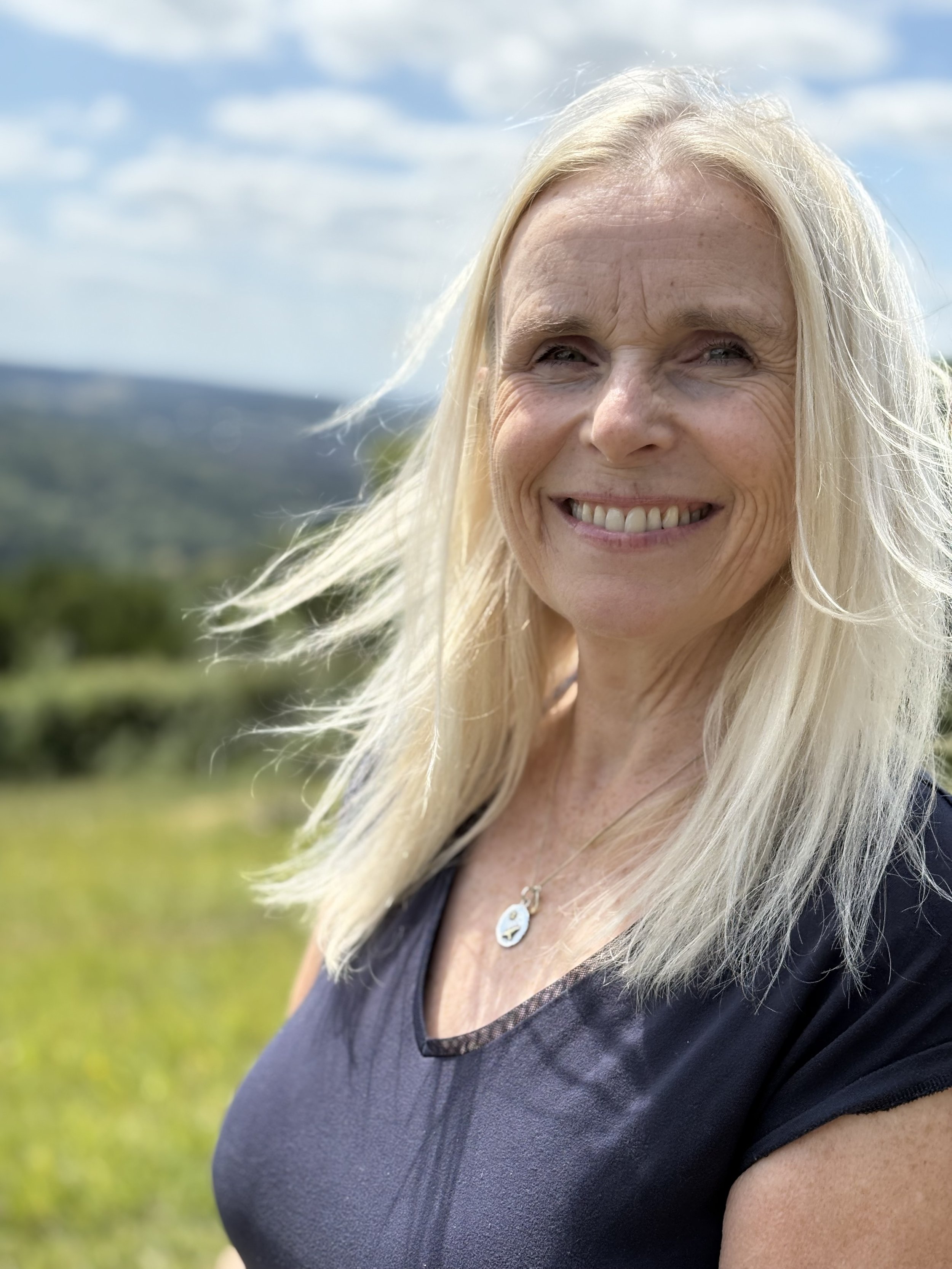 Close-up of a smiling blonde woman outdoors on a sunny day with a grassy field and blue sky in the background.