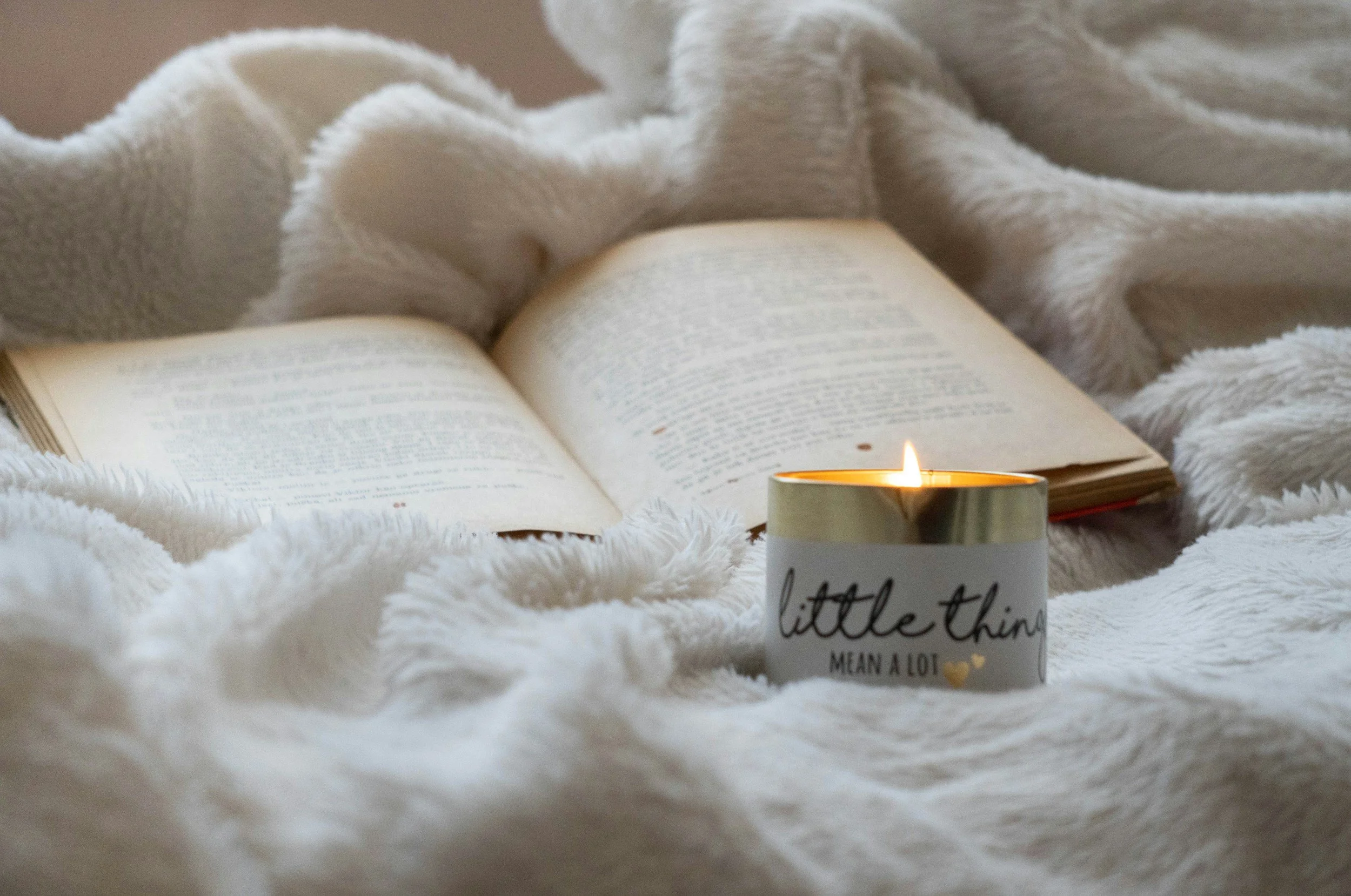 Open book resting on a soft, white, fluffy blanket with a lit candle in a white container that reads 'little things mean a lot' nearby.