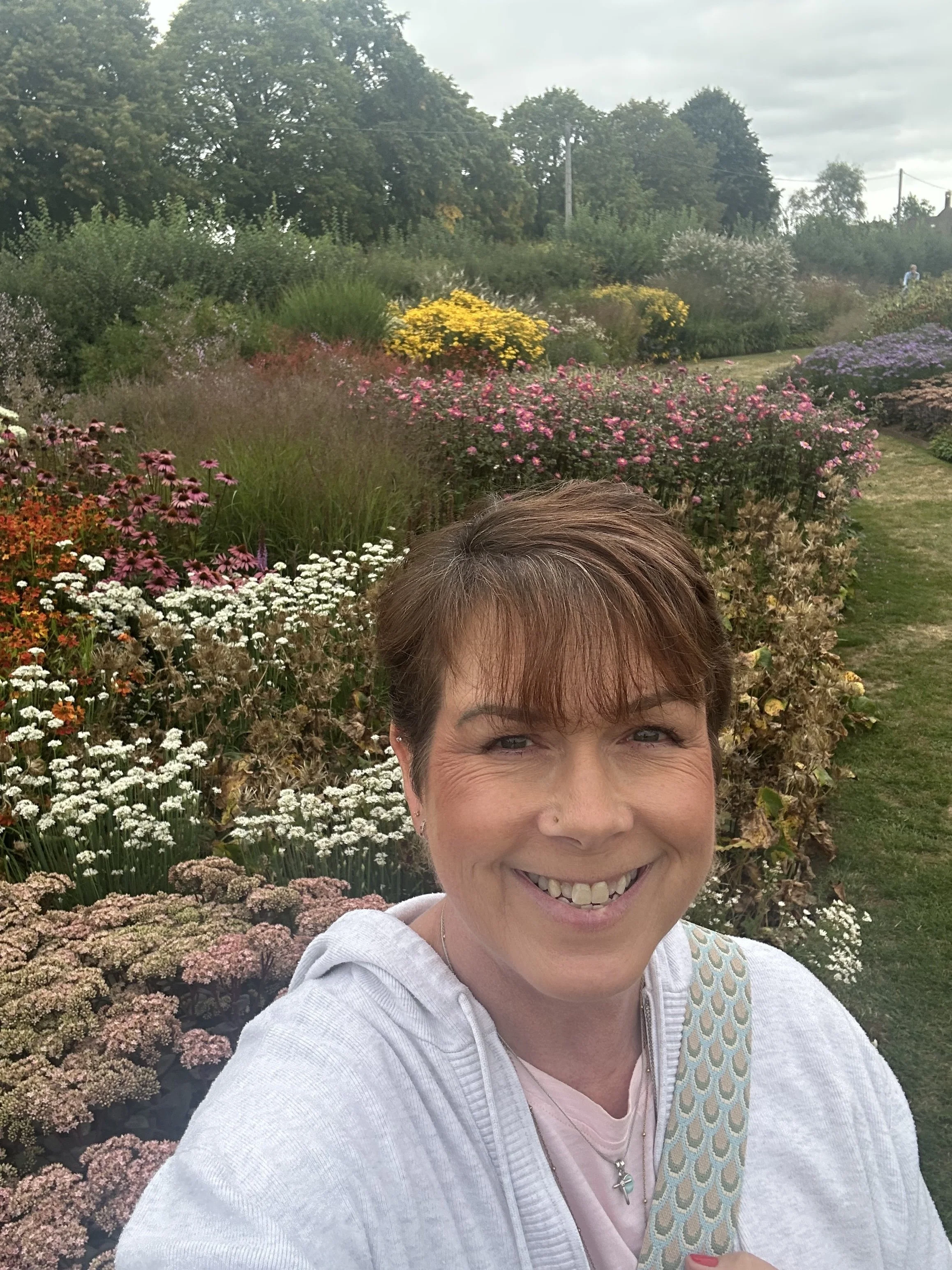 A woman smiling and taking a selfie in a garden filled with colorful blooming flowers.