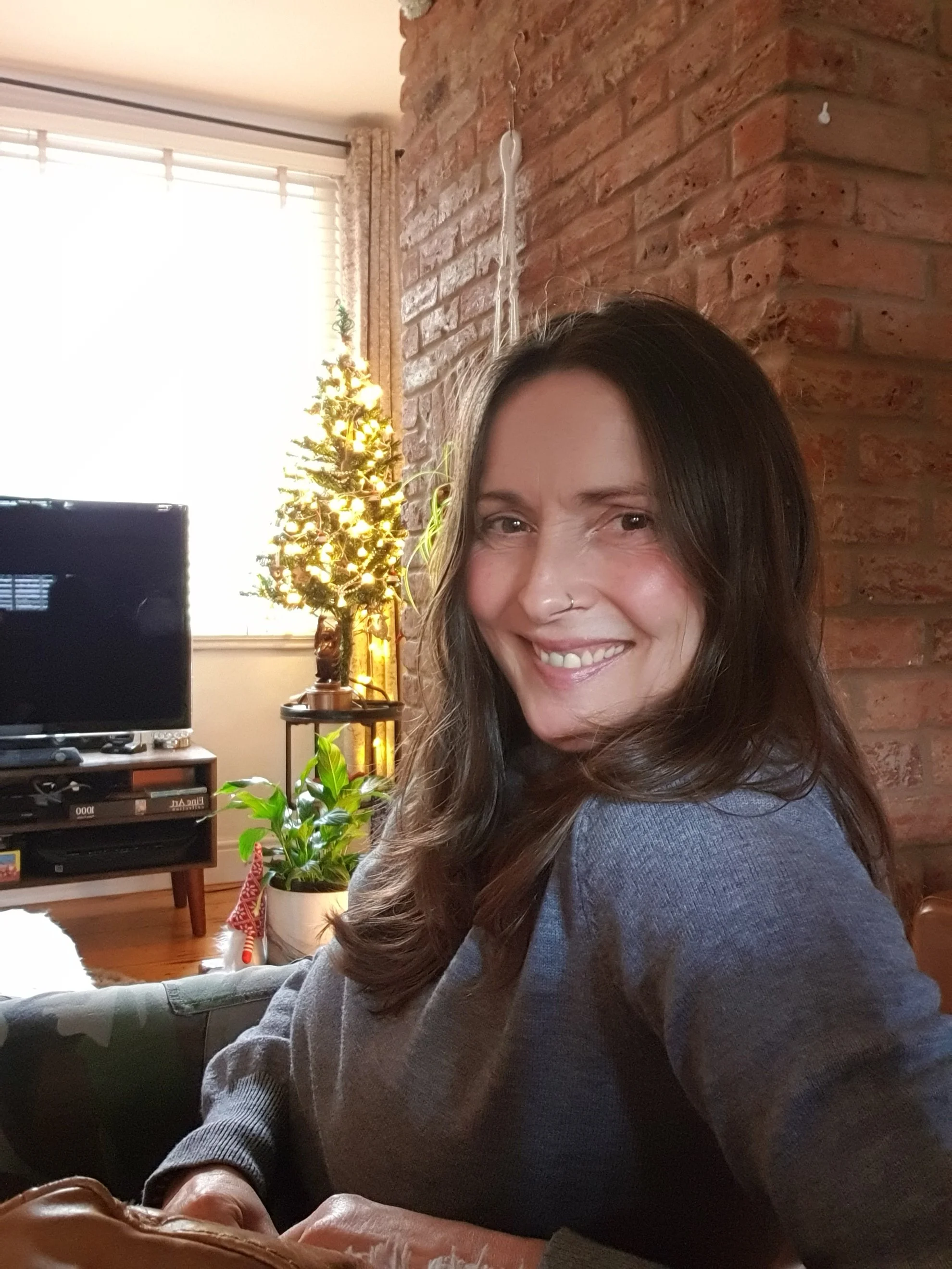 A woman with long brown hair, smiling at the camera in a living room with brick wall, Christmas tree, TV, and plants.