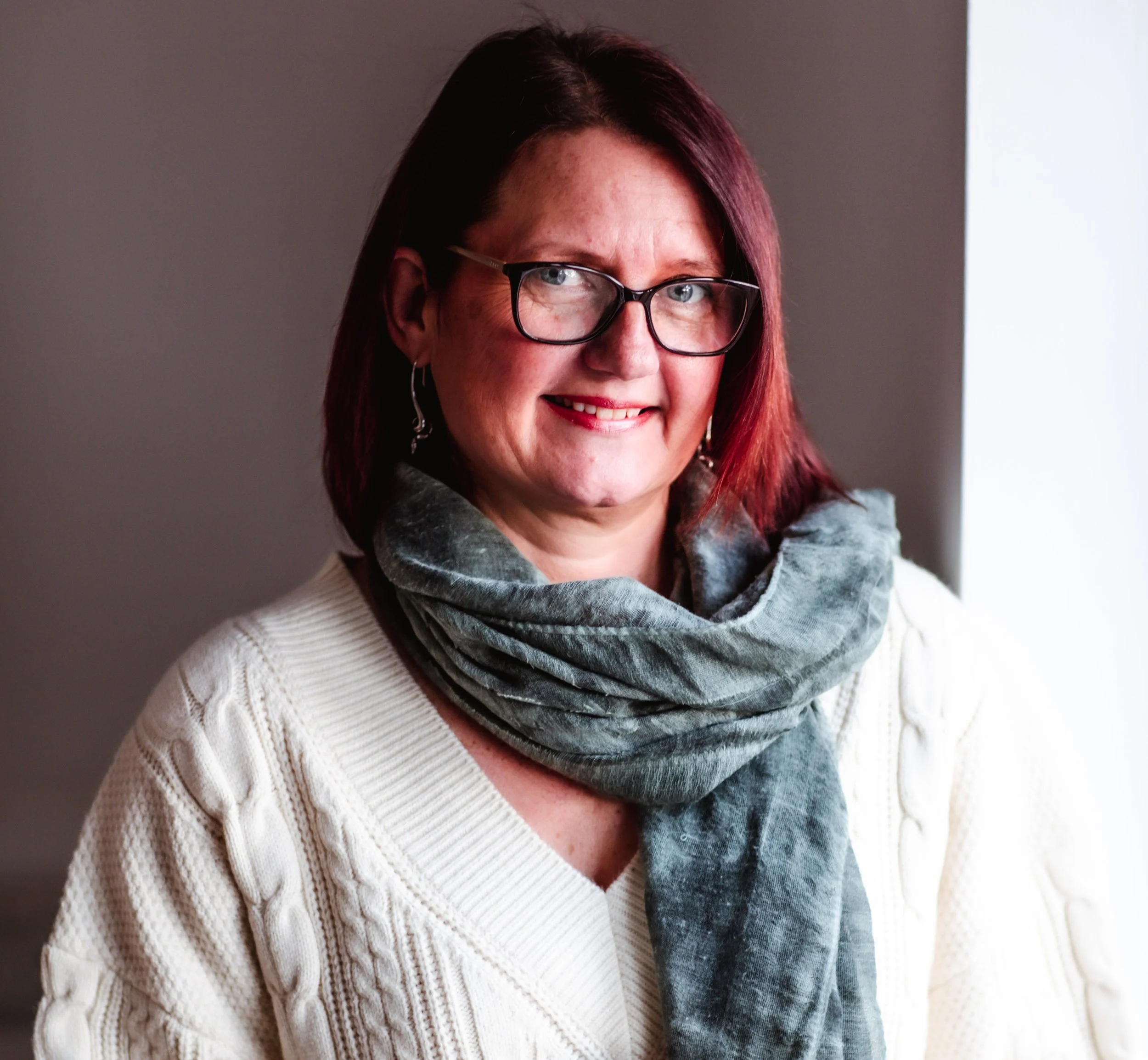 A woman with shoulder-length dark red hair wearing glasses, a cream cable-knit sweater, and a gray scarf, smiling and standing near a window.