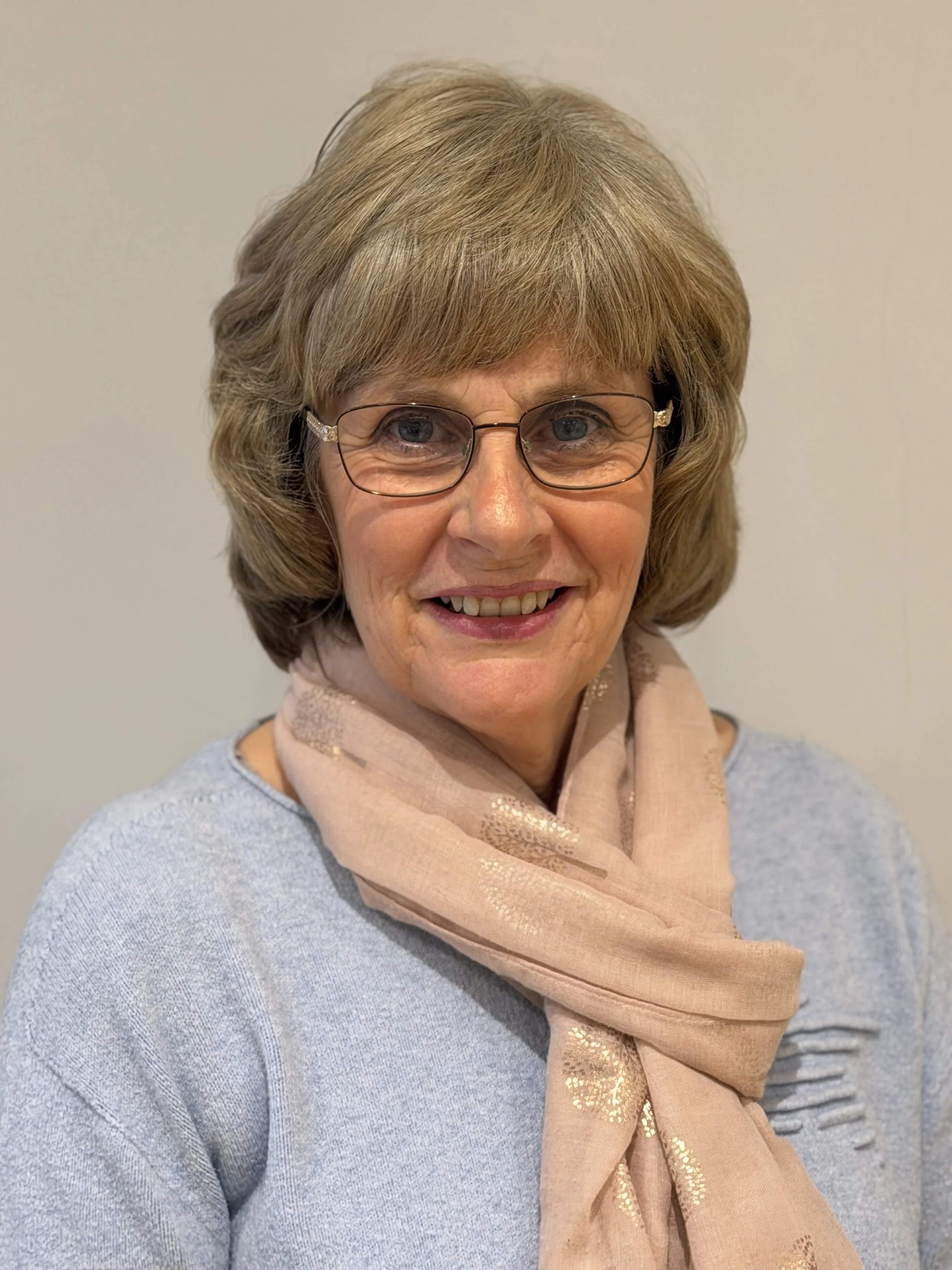 A smiling woman with glasses, short blonde hair, wearing a light gray sweater and a pink scarf with metallic gold accents, against a plain light-colored background.