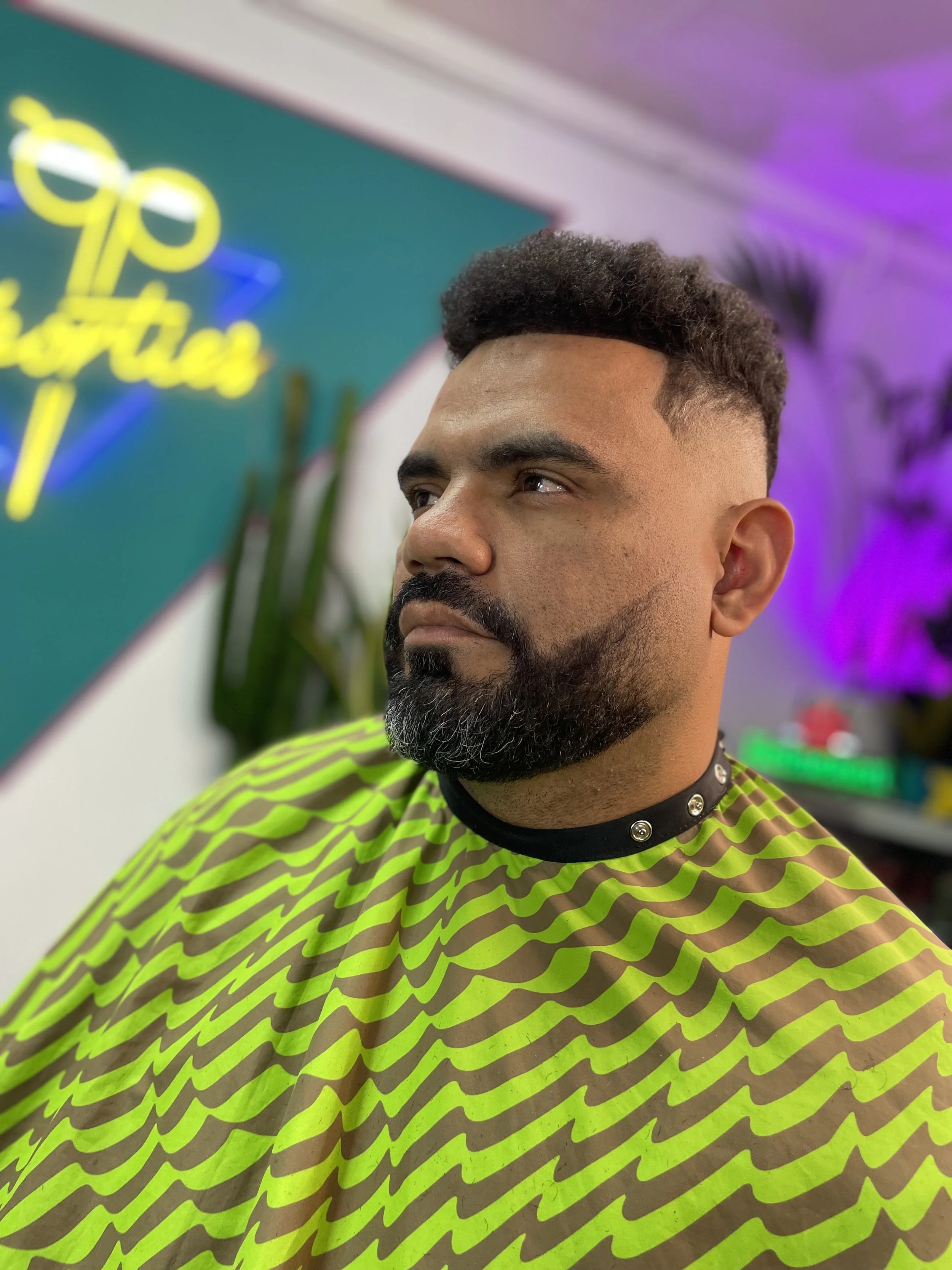 A man with dark hair and a beard, wearing a neon green and black patterned barber cape and a black studded collar, sitting in a barber shop with colorful lights and decor in the background.