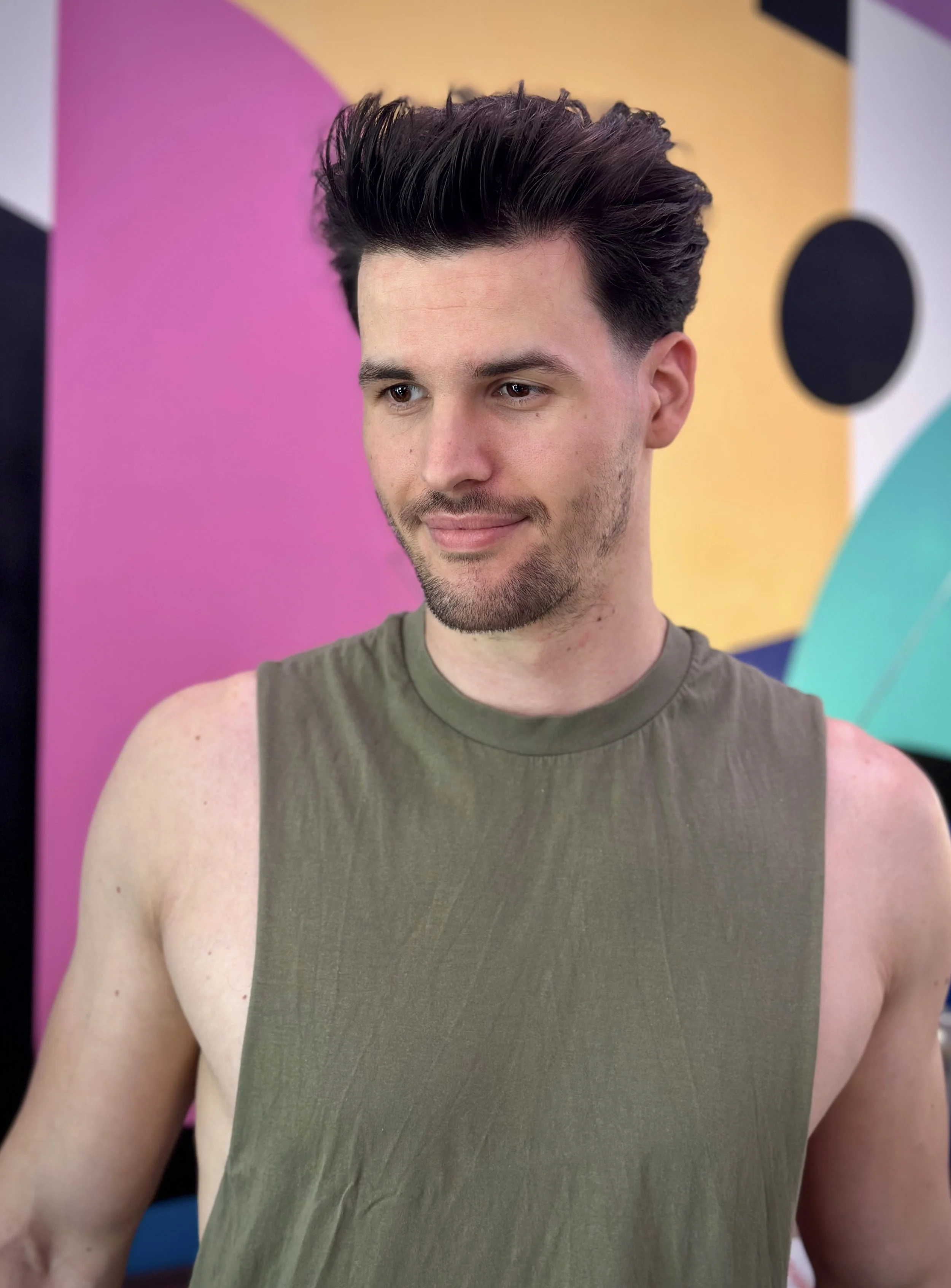 A young man with dark, styled hair and facial stubble, wearing a sleeveless olive green shirt, standing in front of a colorful abstract background.
