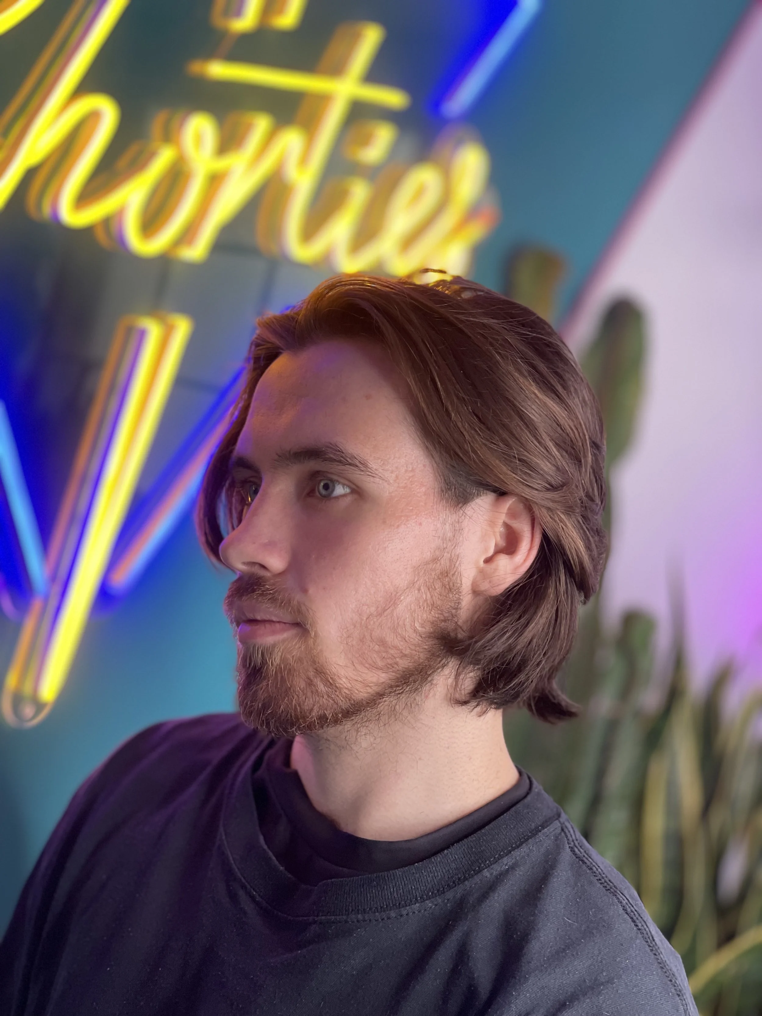 Side profile of a young man with medium-length brown hair, light beard, wearing a black shirt, standing in front of a neon sign with yellow and blue lights that reads 'thrive', with green plants visible in the background.