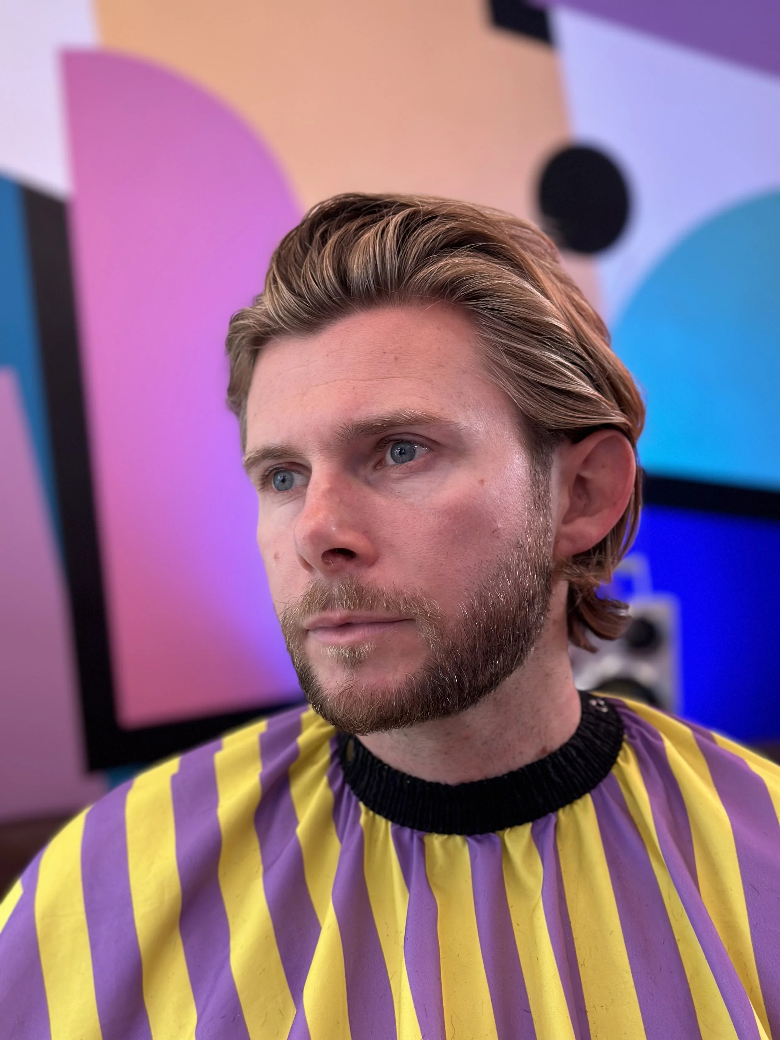 A man with a beard and light brown hair, wearing a yellow and purple striped cape, is sitting in a colorful room with abstract wall art in pink, blue, and purple.