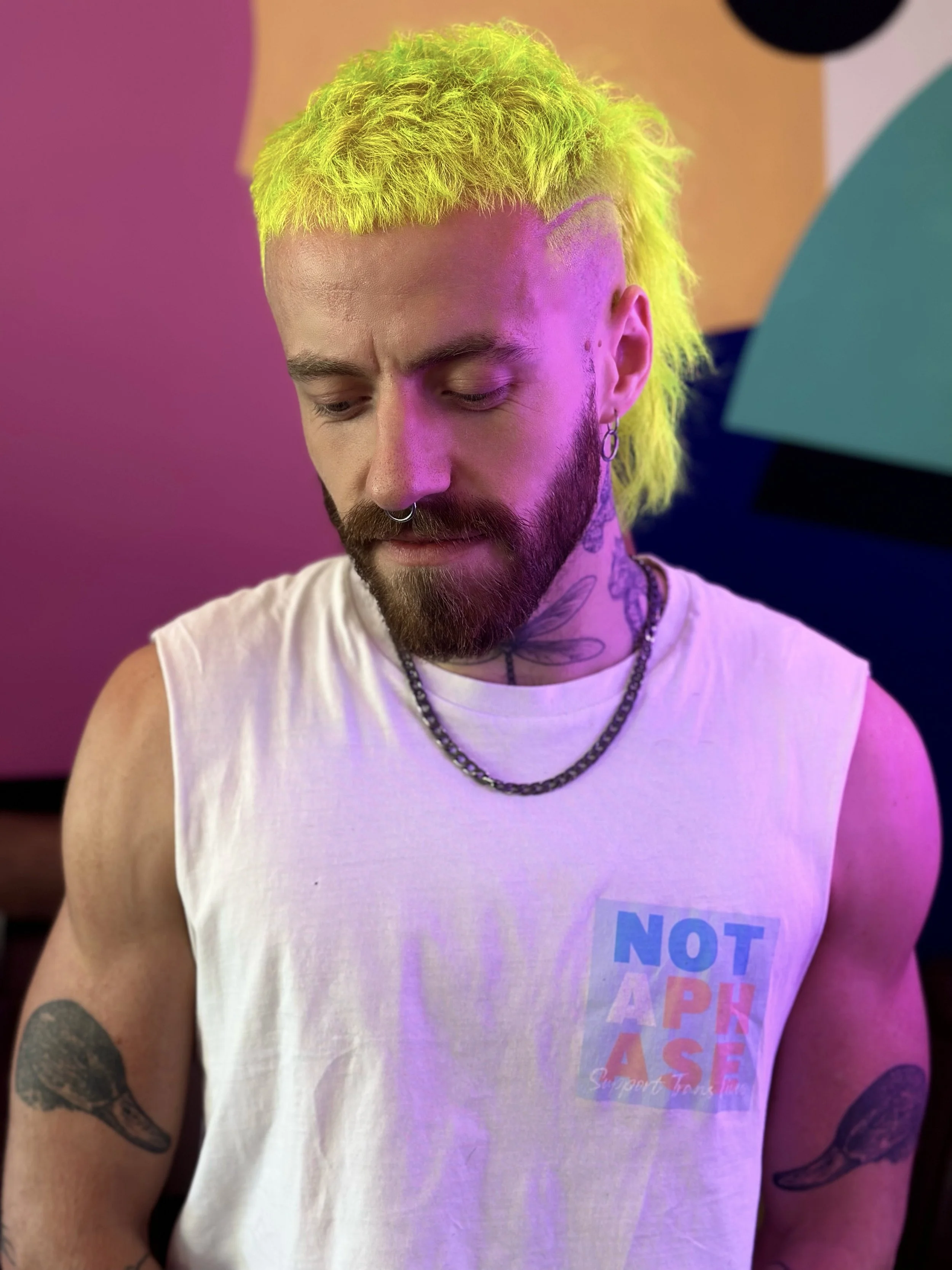 A young man with yellow-green hair, tattoos, and piercings, wearing a sleeveless white shirt with a colorful logo, standing against a vibrant, abstract background.