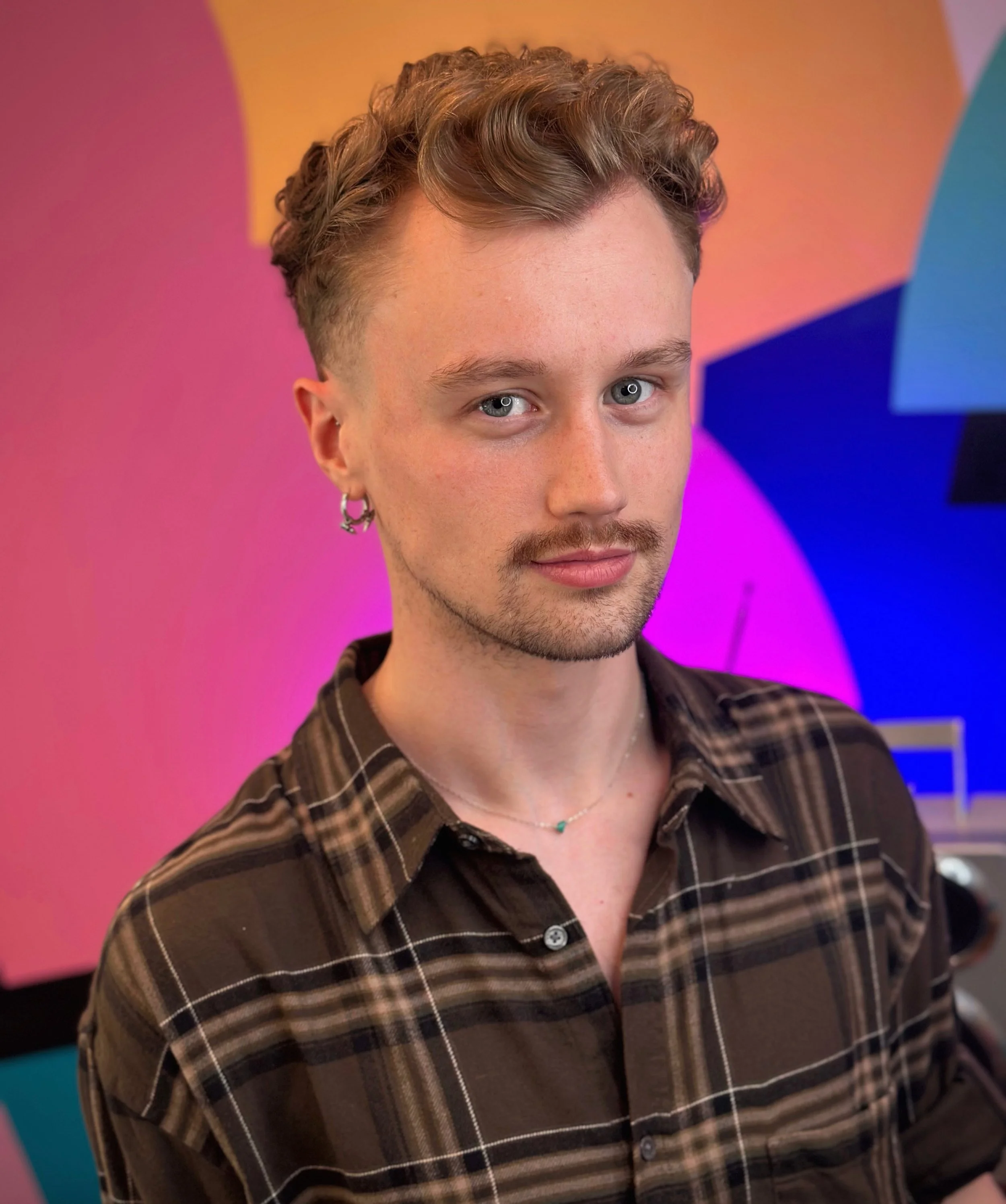 A young man with styled brown hair, wearing a plaid shirt, earrings, and a necklace, standing in front of a colorful abstract background with pink, orange, blue, and purple shades.
