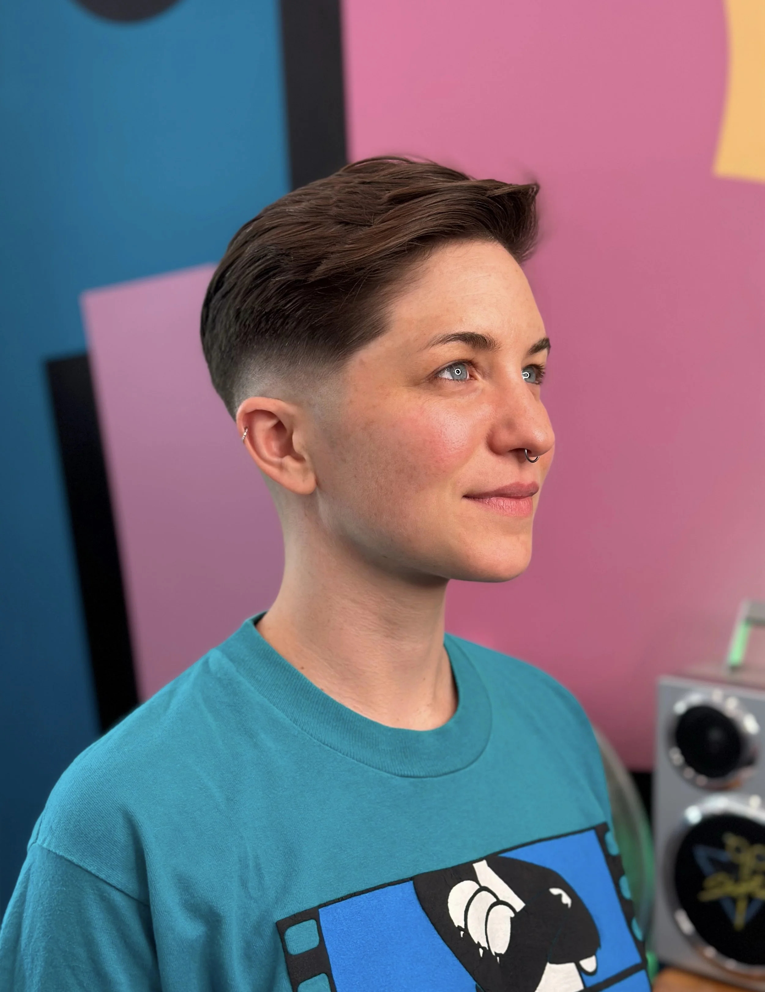 A young woman with short, styled brown hair, blue eyes, and a septum piercing, smiling and looking slightly to the right. She is wearing a turquoise t-shirt with a digital art graphic on it. The background features colorful pastel panels in pink, pur