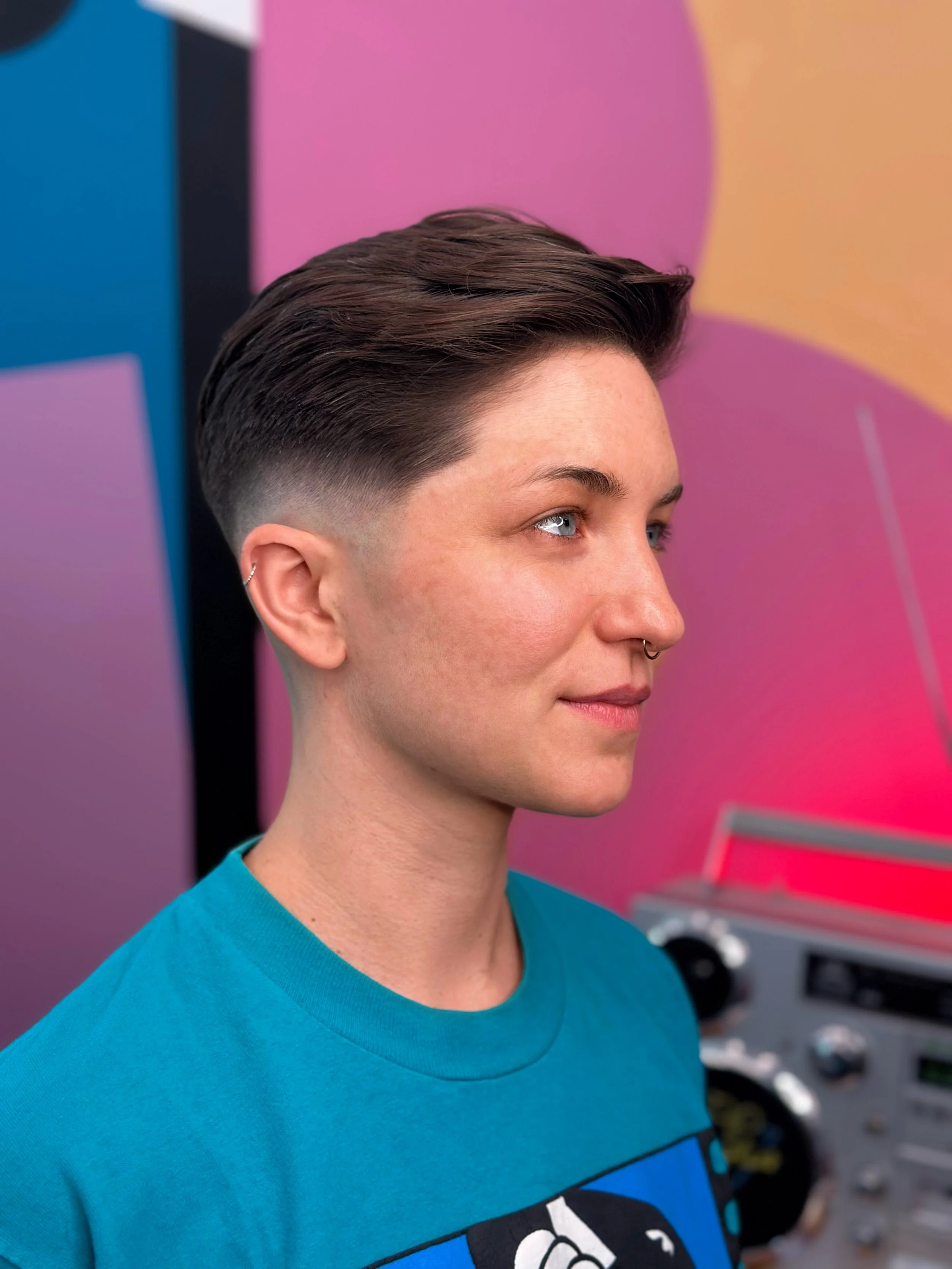 Person with short hair styled in a fade, wearing a blue shirt, standing in front of a colorful, abstract wall with a pink, purple, and yellow background, and electronic equipment in the background.