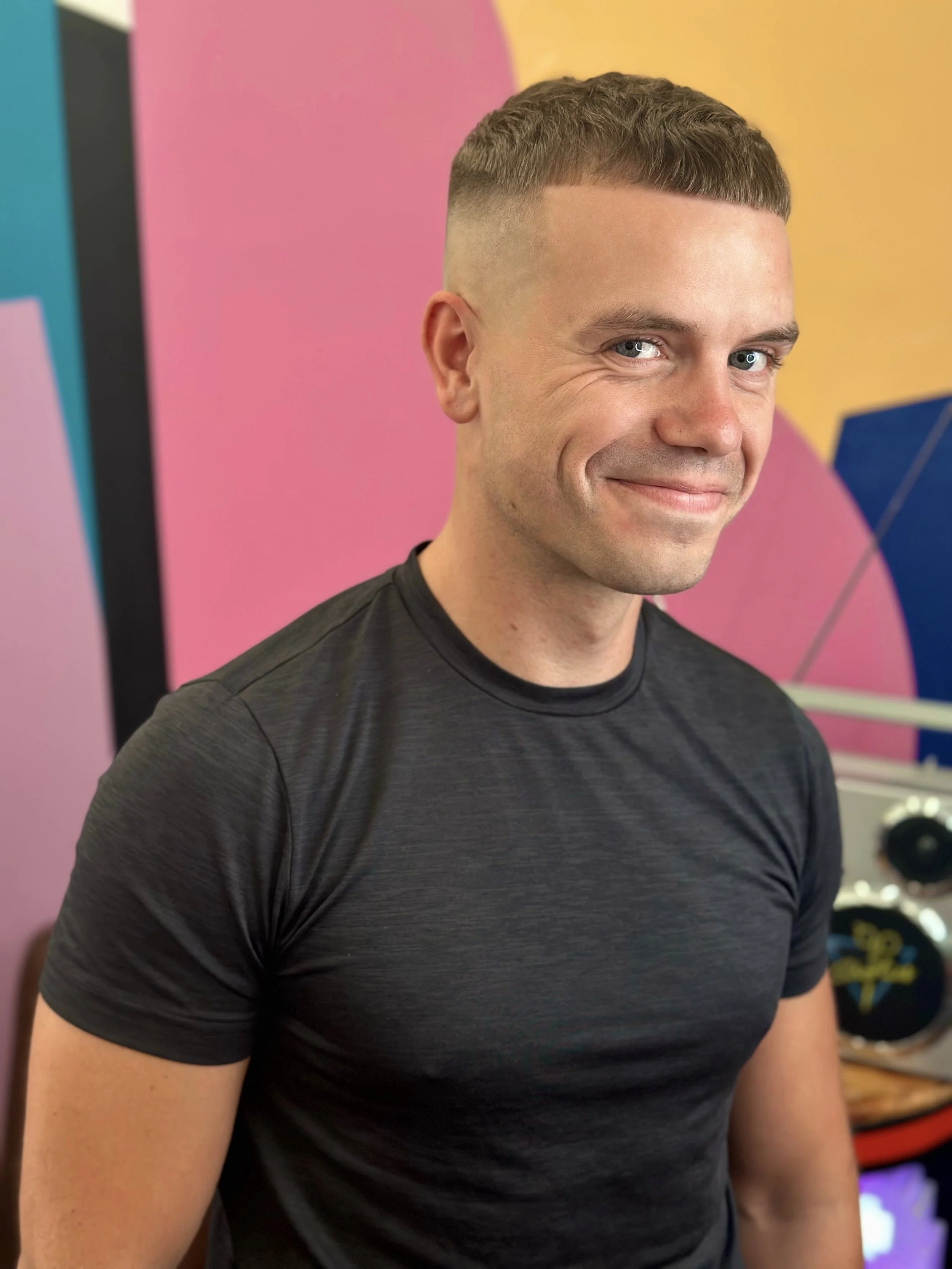 A man with short brown hair and blue eyes is smiling at the camera, wearing a black fitted T-shirt, standing in front of a colorful, geometric background.