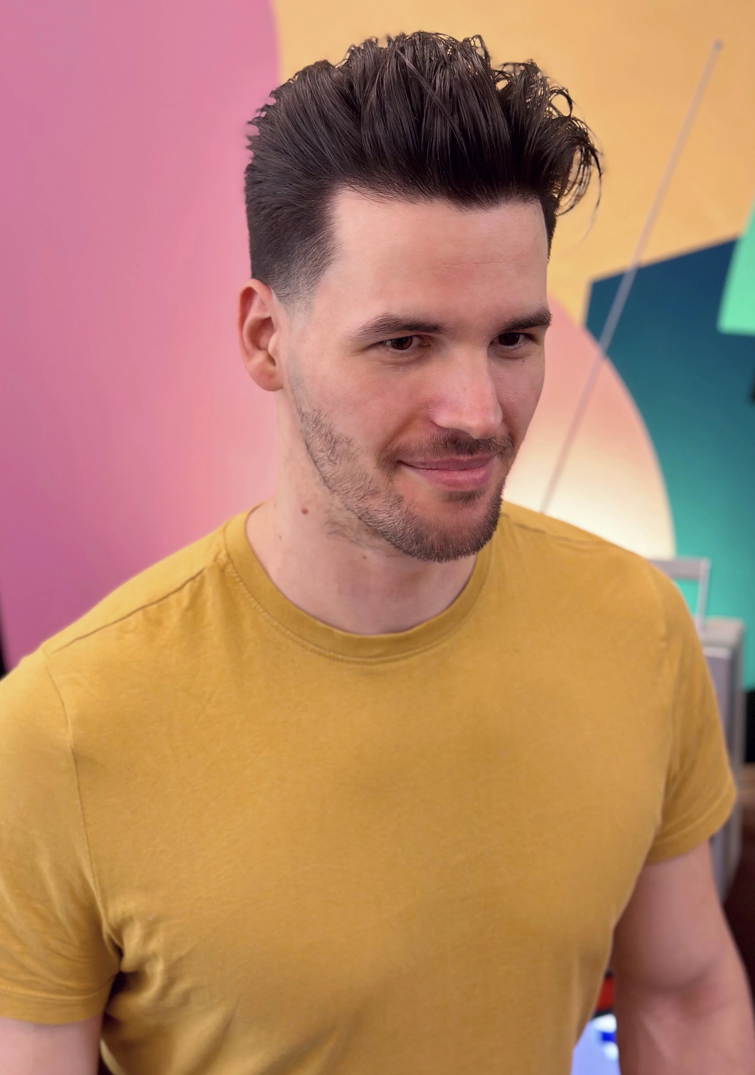 A young man with dark, styled hair and a trimmed beard wearing a yellow t-shirt, standing in front of a colorful, abstract background with pink, yellow, and teal shapes.