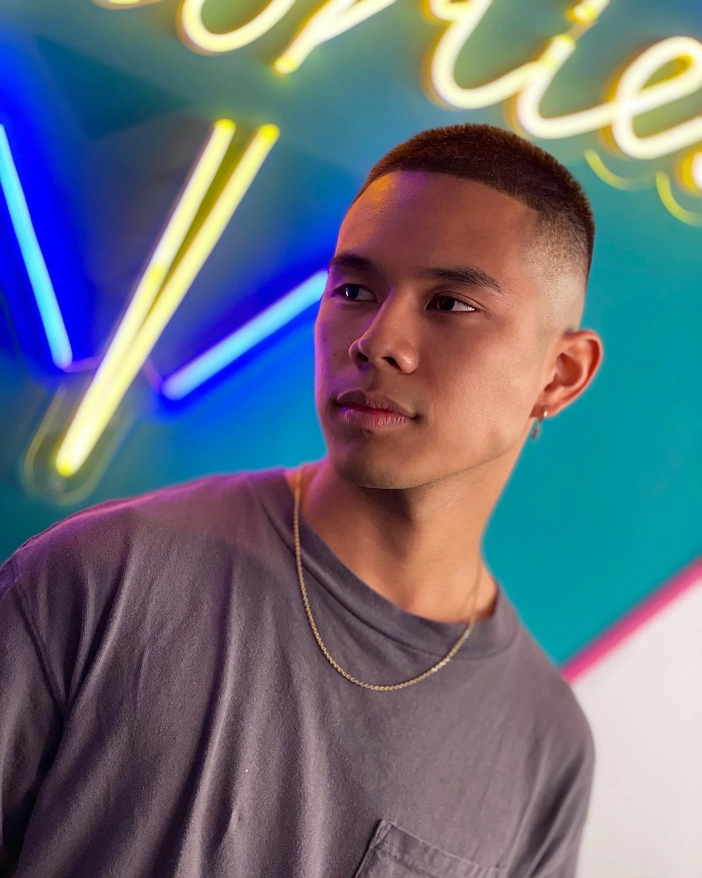 Young man with short hair and pierced ear wearing a grey t-shirt and gold chain, standing in front of a neon sign with colorful lights.