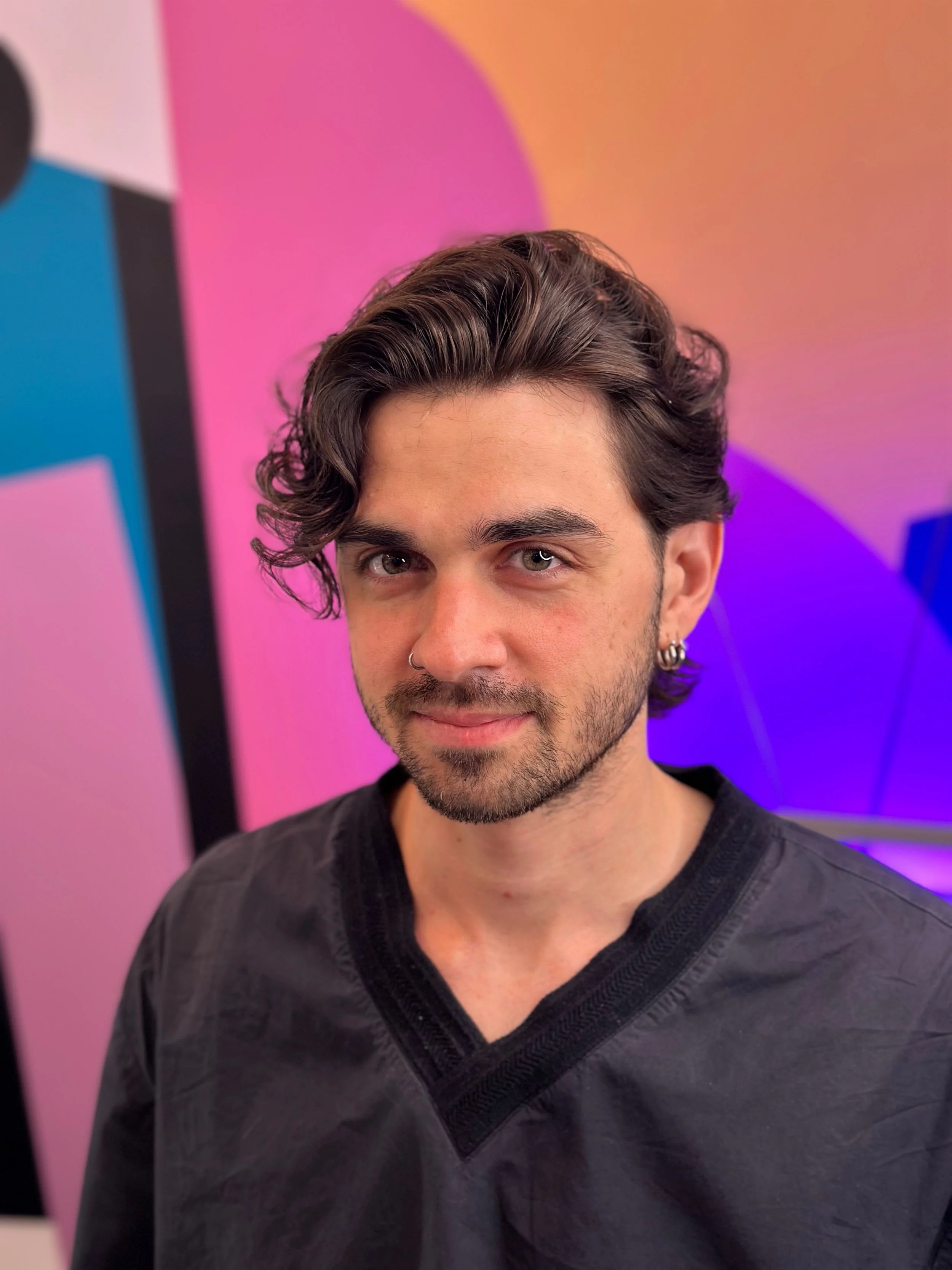A young man with wavy dark hair, a beard, and piercings on his nose and ears, wearing a black shirt, standing in front of a colorful abstract background with pink, purple, and orange hues.