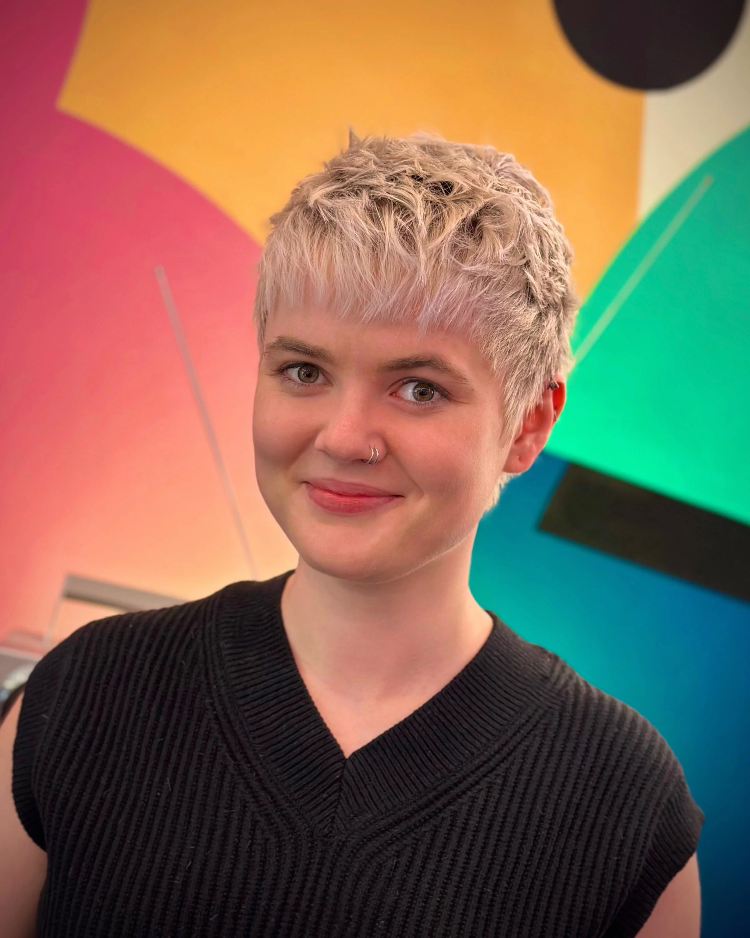 A young woman with short blonde hair, fair skin, and a nose ring smiling in front of a colorful abstract background.