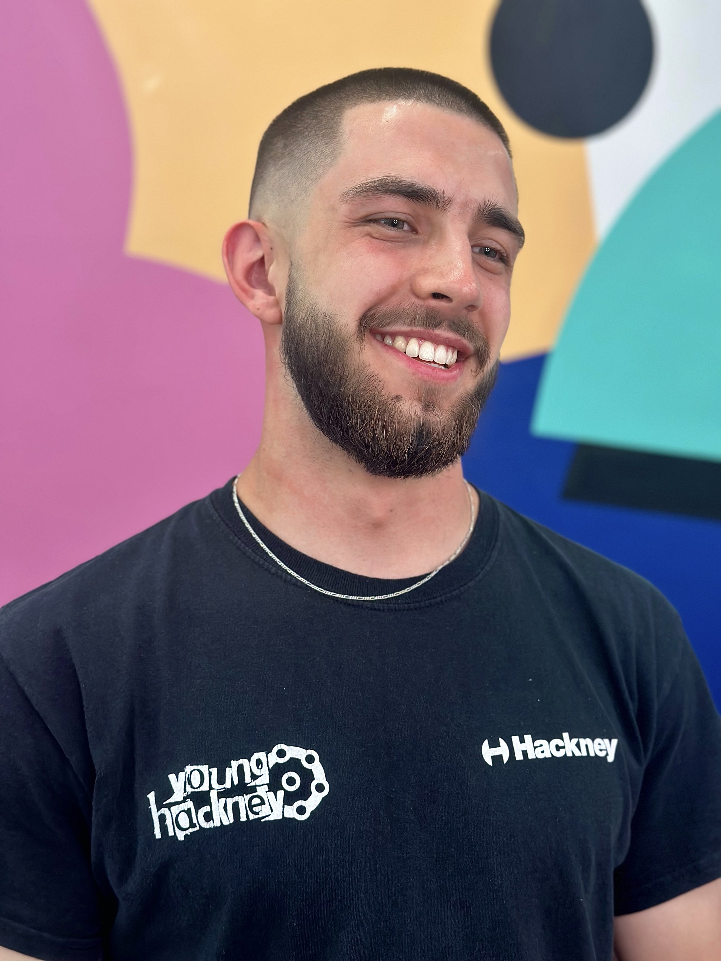 A smiling young man with a beard and short hair wearing a black Hackney t-shirt with a project logo and a silver necklace, standing in front of a colorful abstract background.