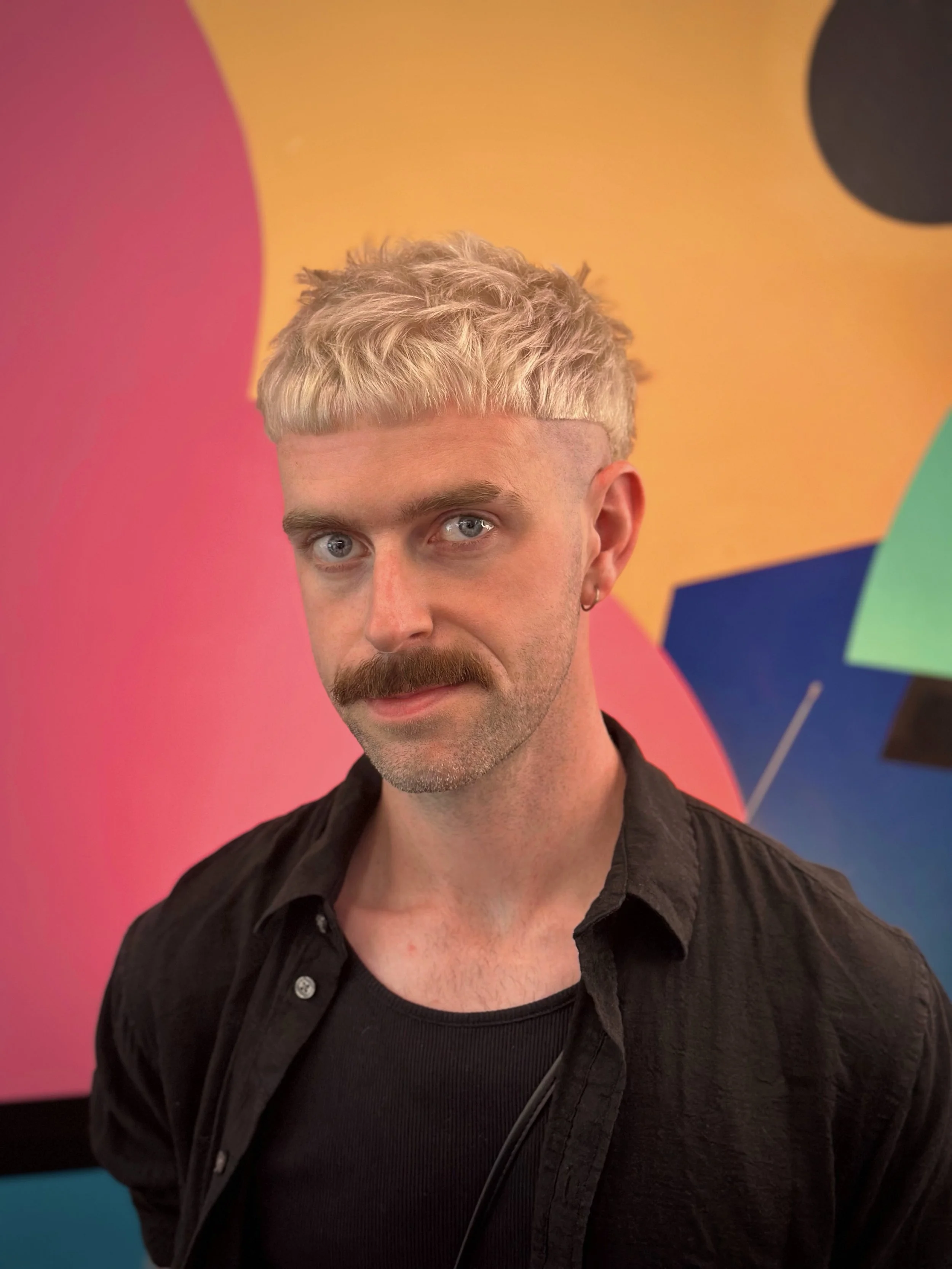 A man with pale blonde hair and a mustache, wearing a black shirt, stands in front of a colorful geometric background.