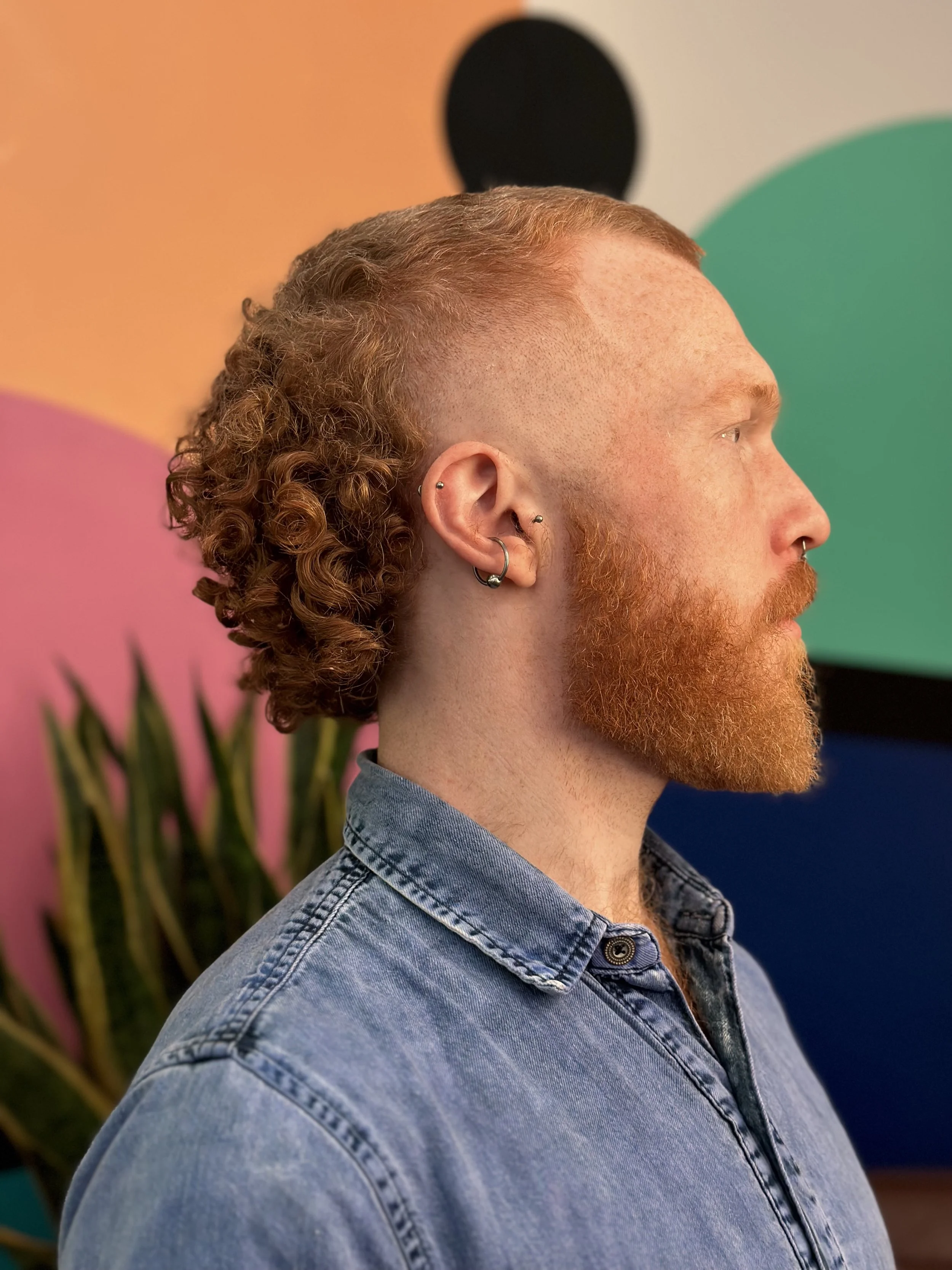 Profile side of a man with red hair and beard, wearing a denim shirt, and multiple earrings, standing in front of a colorful abstract background.
