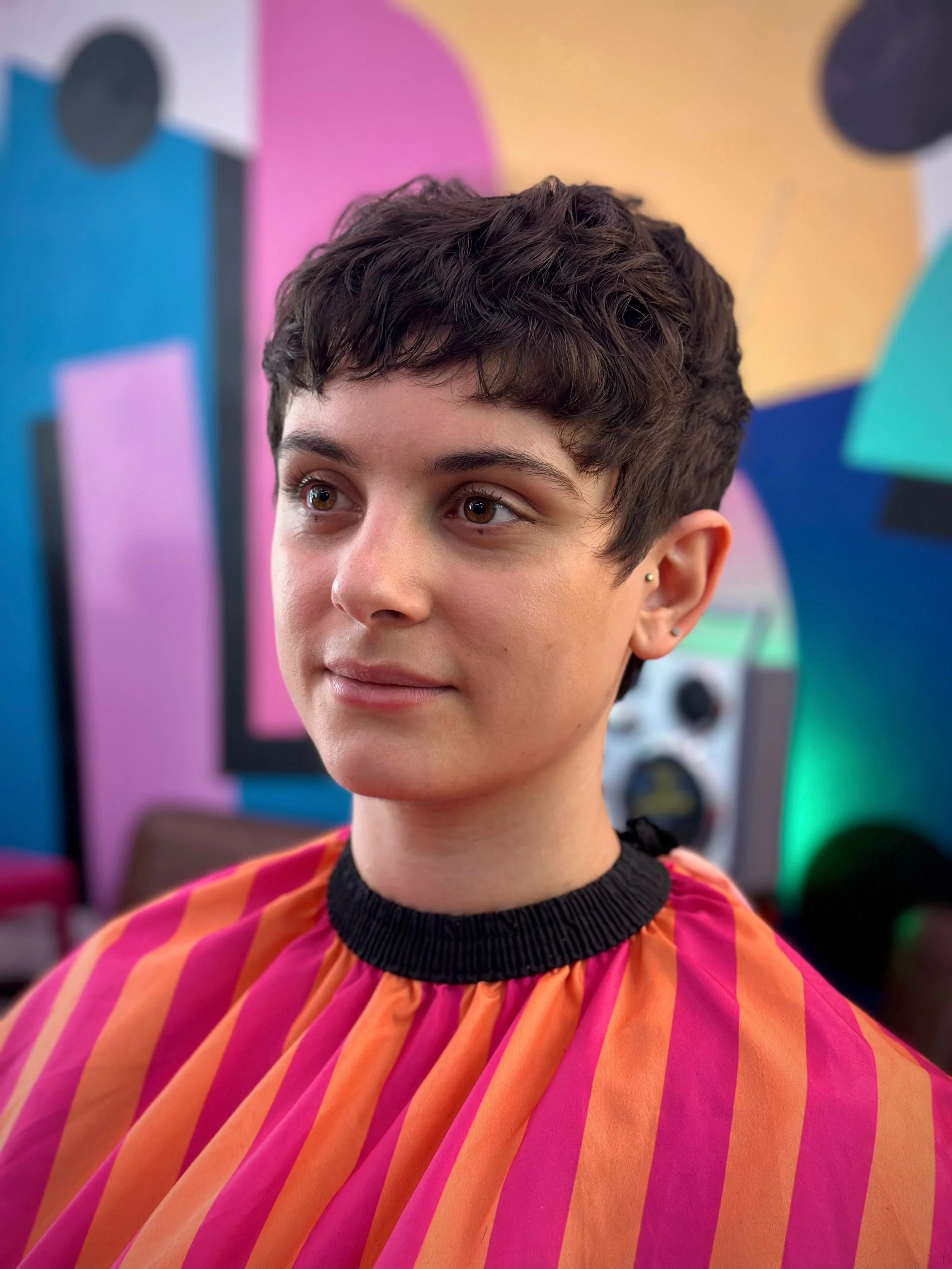 A young person with short, dark hair looking slightly to the side, wearing a colorful striped cape in pink and orange with a black neckline, in a room with vibrant abstract wall art and a speaker in the background.