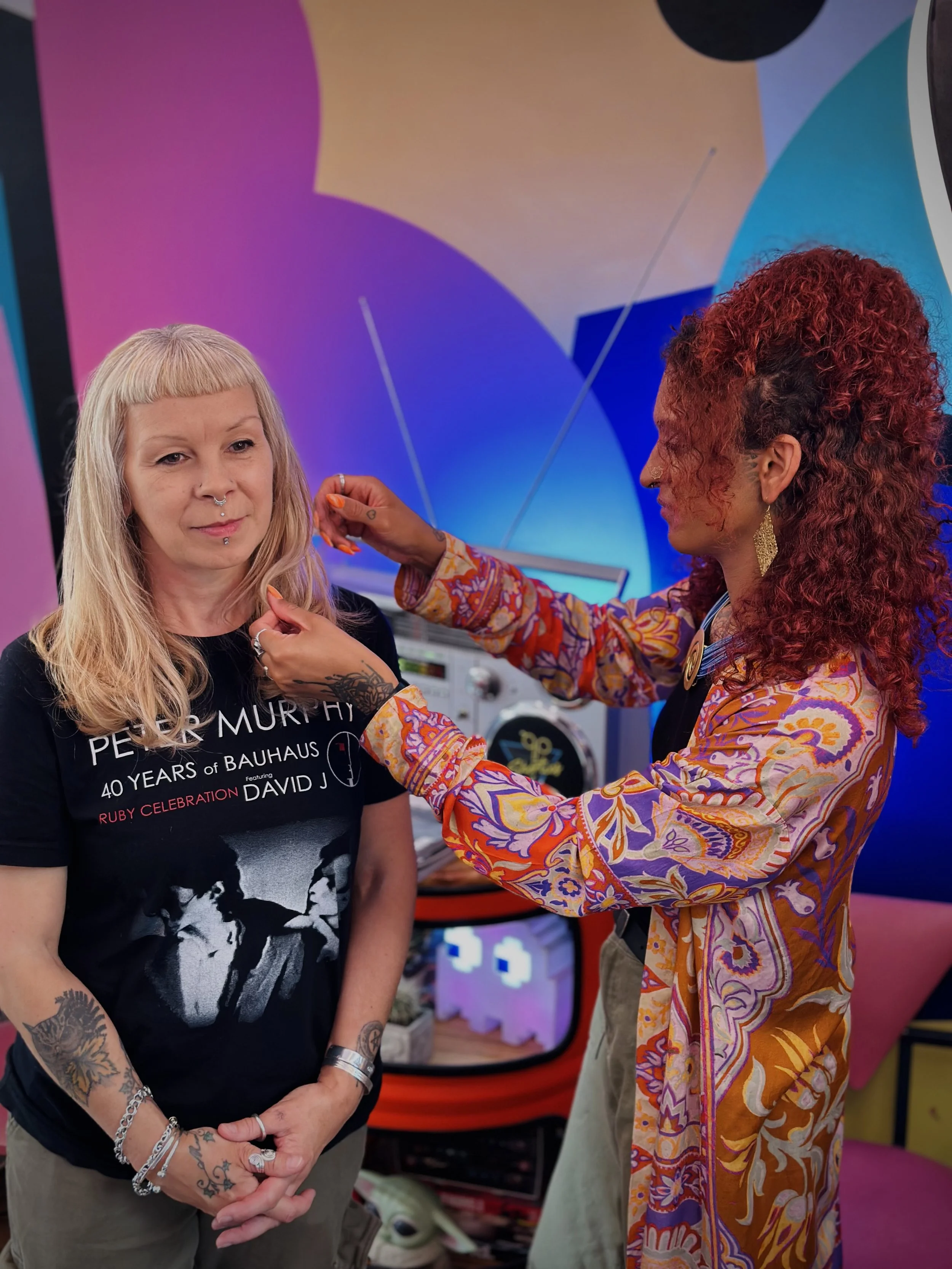 Two women, one with long blonde hair and tattoos, and the other with curly red hair, are in a vibrant, colorful room. The woman with red hair is applying makeup or jewelry to the woman with blonde hair, who is wearing a black T-shirt with graphic tex
