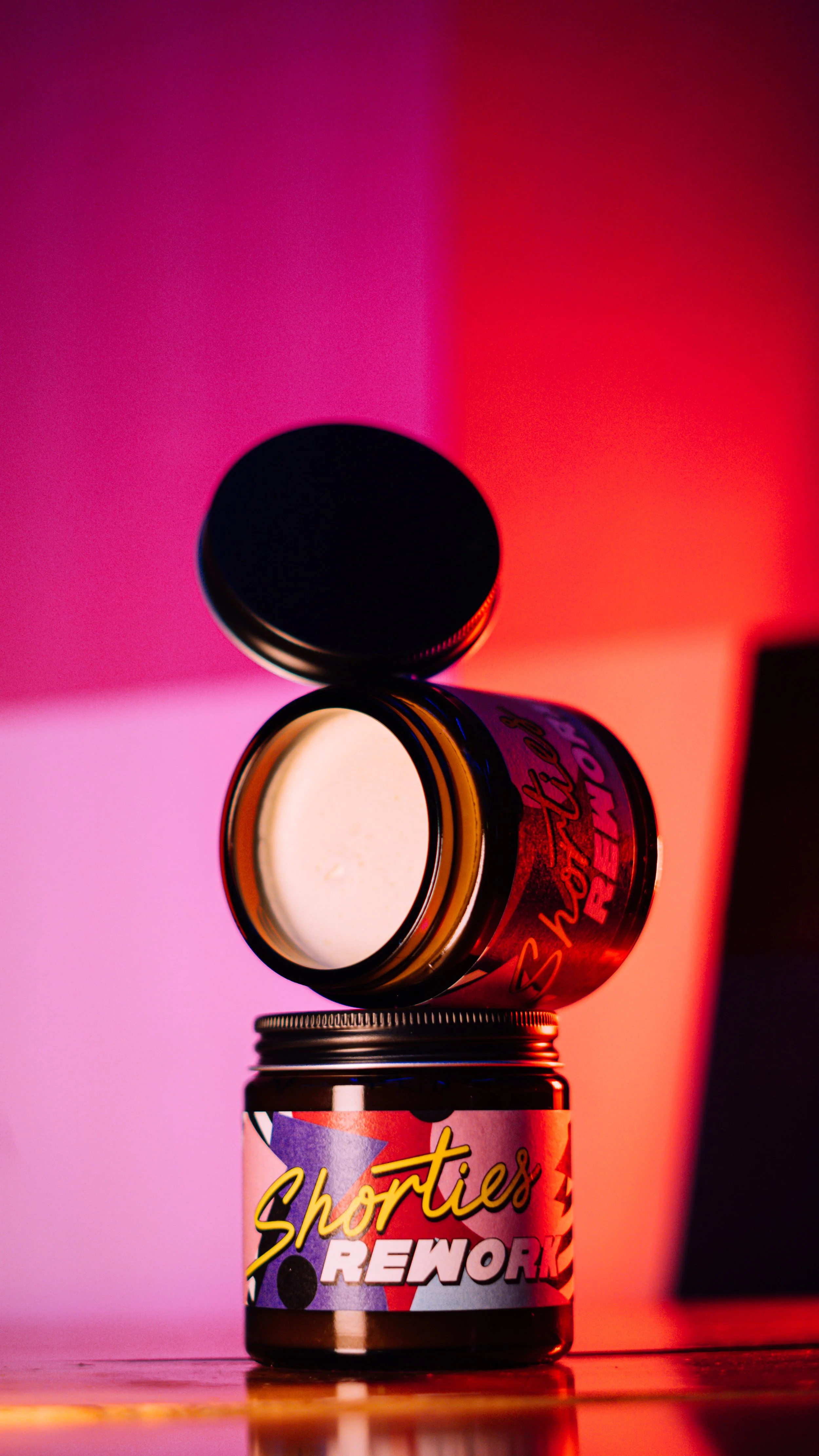 Stacked jars with colorful labels and a black lid, set against pink and red backlighting.