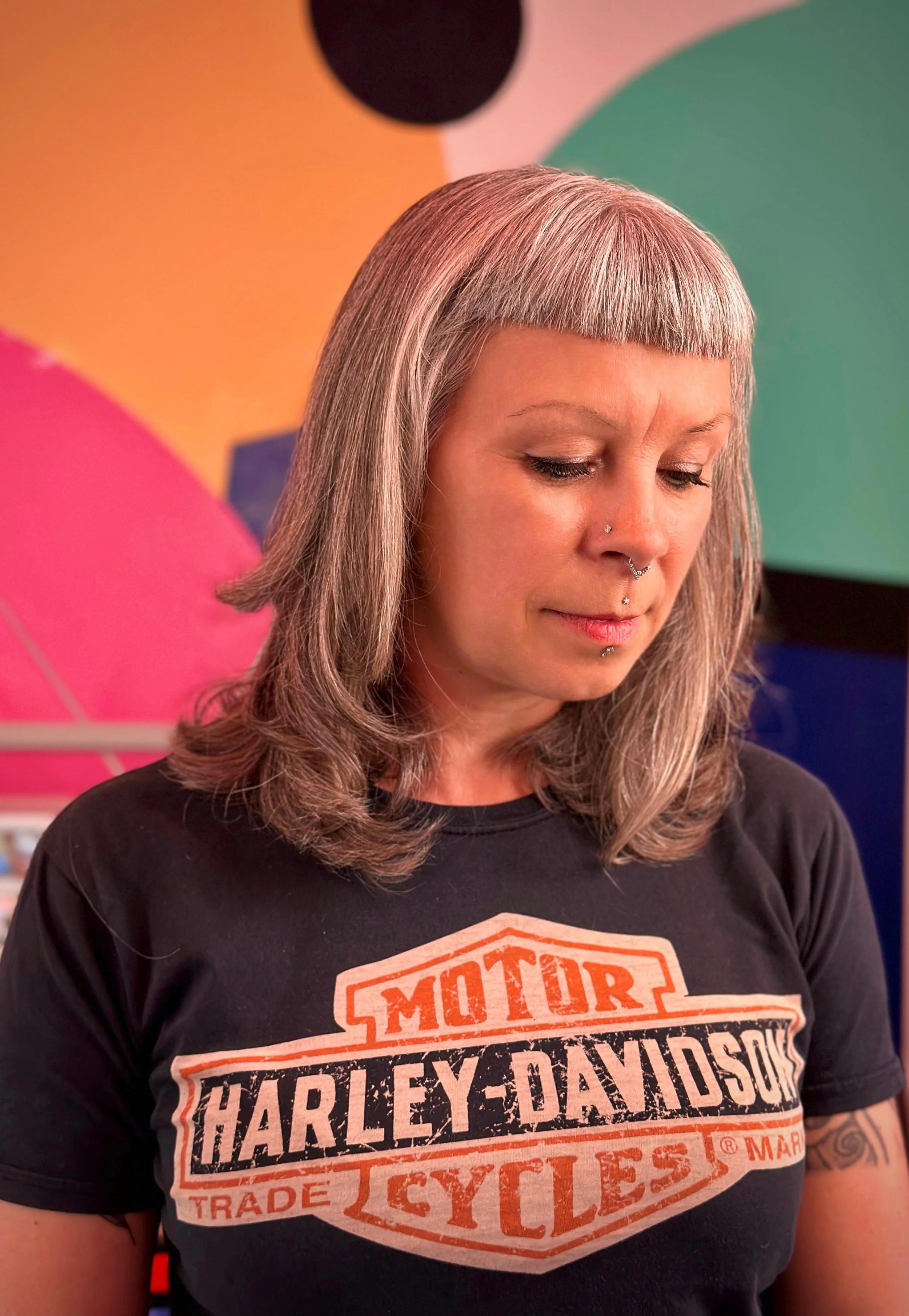 A woman with silver hair and multiple facial piercings wears a black Harley-Davidson t-shirt.