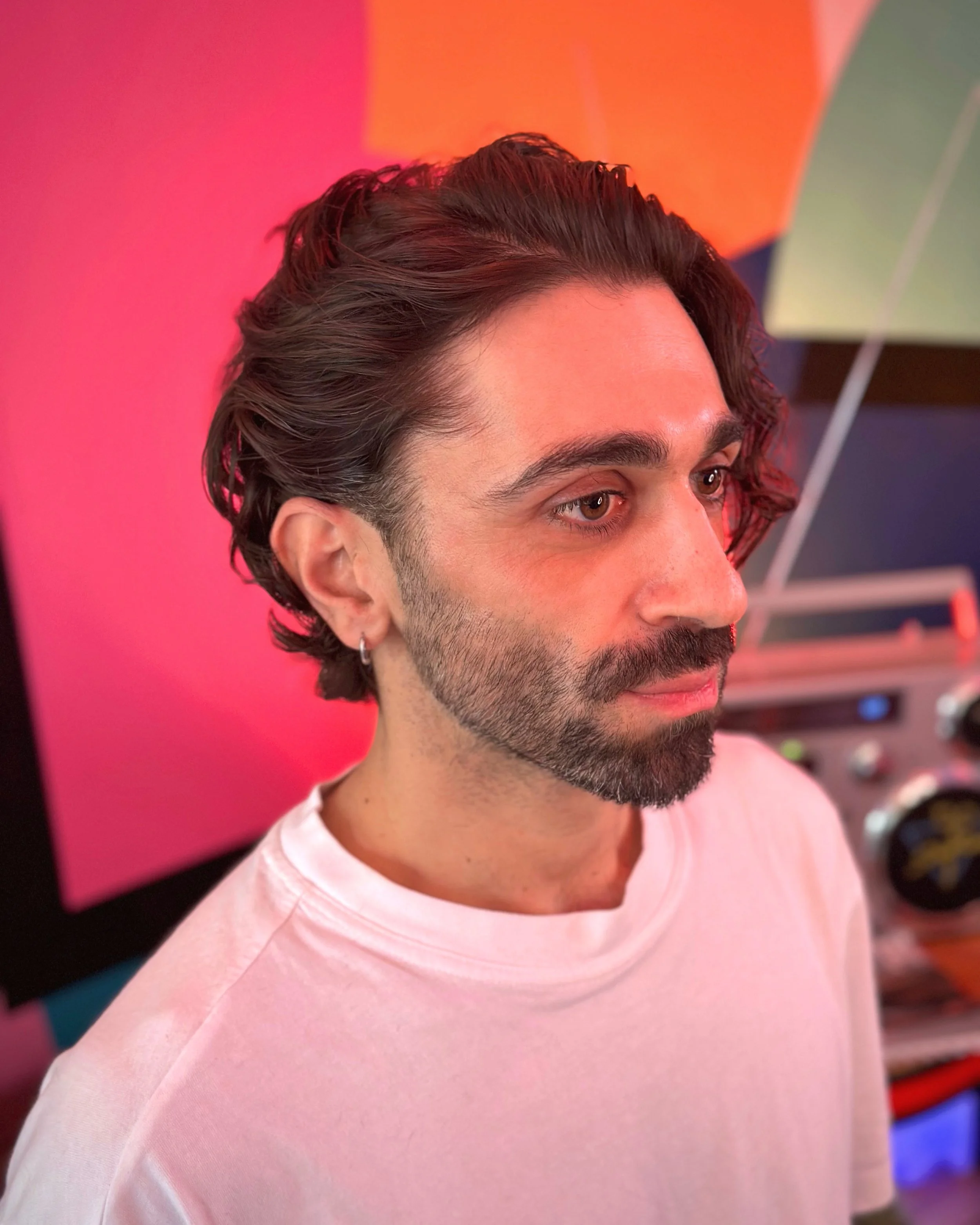 A man with dark, curly hair, a beard, and wearing a white shirt, standing in front of colorful geometric background, looking to the right.