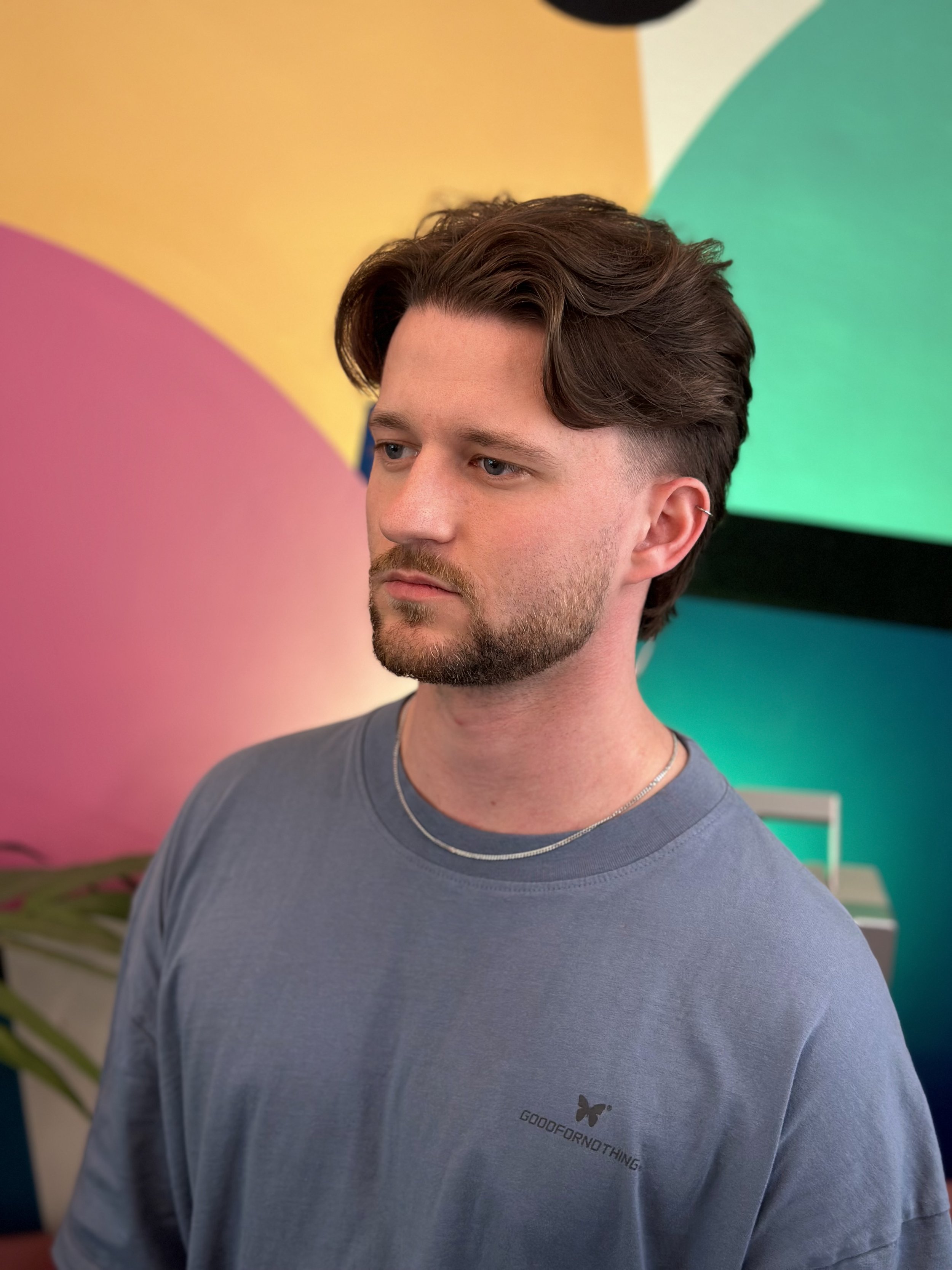 A young man with brown hair, a beard, and blue eyes looking slightly to the left, wearing a gray t-shirt with a small butterfly logo and the word 'GOODFORNOTHING' on the chest, standing in front of a vibrant, colorful abstract background with hues of