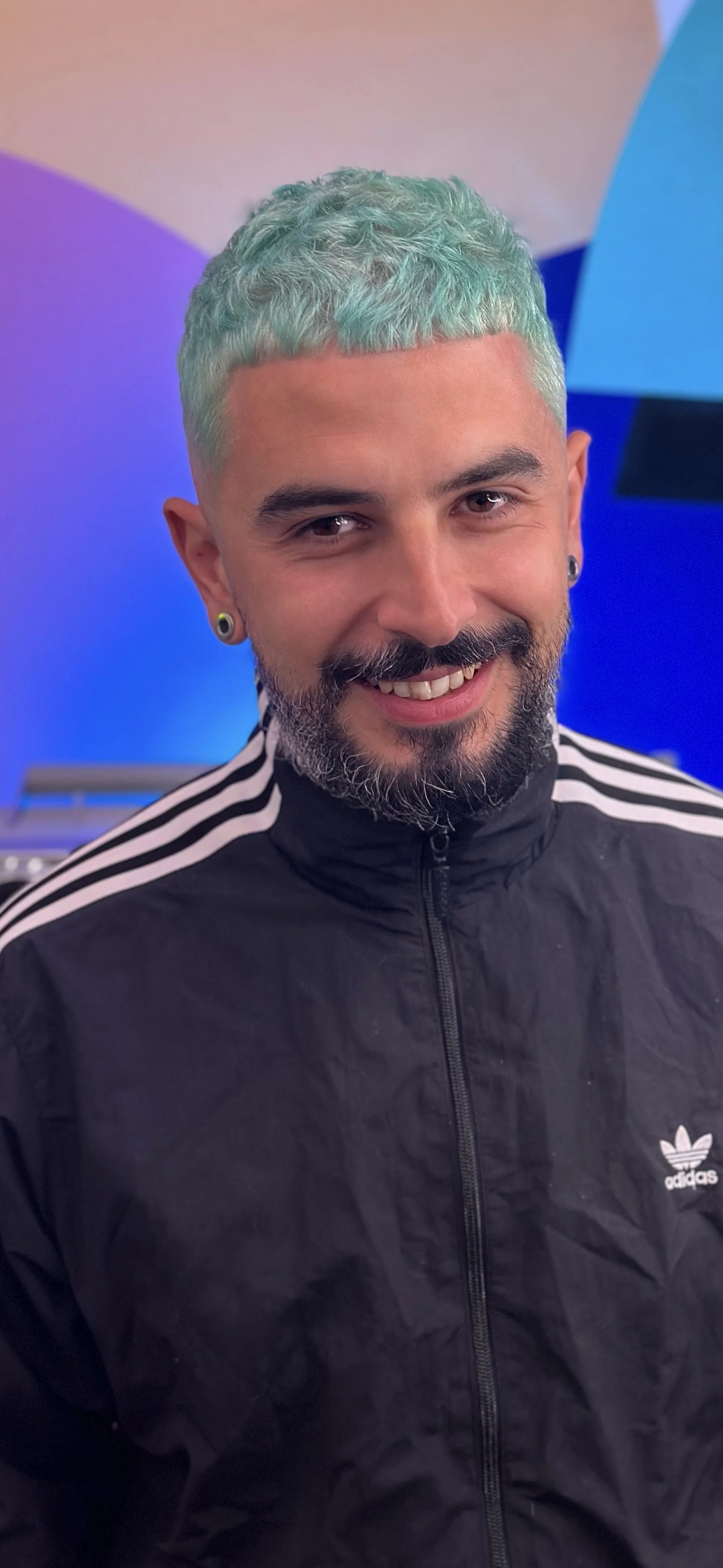 Smiling man with short, curly, pastel green hair, beard, and earrings, wearing a black Adidas jacket with white stripes. Background features colorful, abstract shapes.
