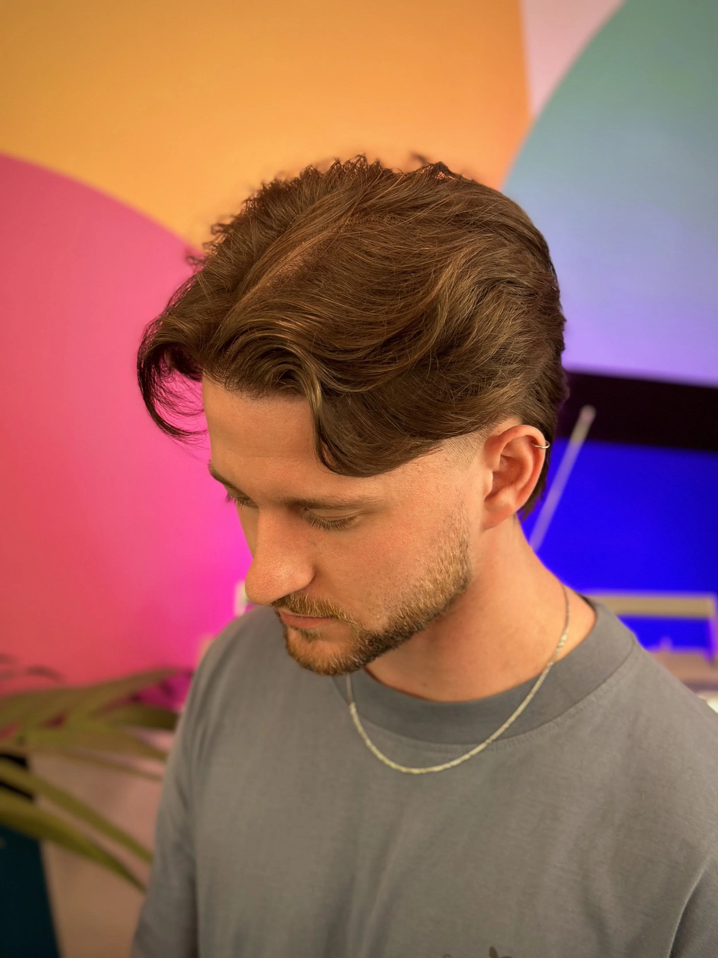 A young man with brown styled hair and a beard, wearing a gray t-shirt and a silver necklace, standing in front of a colorful background with pink, orange, purple, and teal lights.