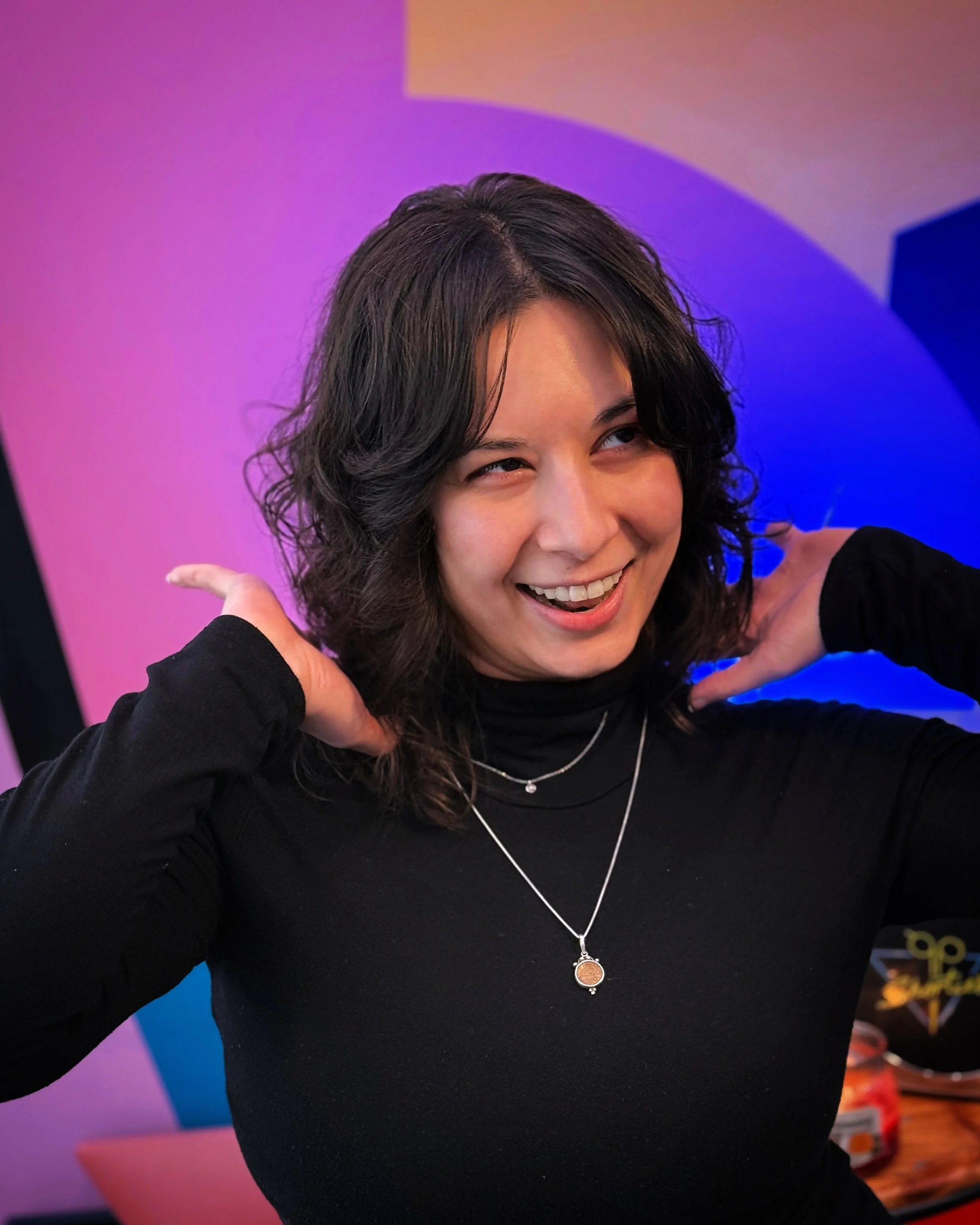A woman with dark wavy hair smiling and touching her neck with her hands, wearing a black top and layered necklaces, with colorful abstract background lighting.