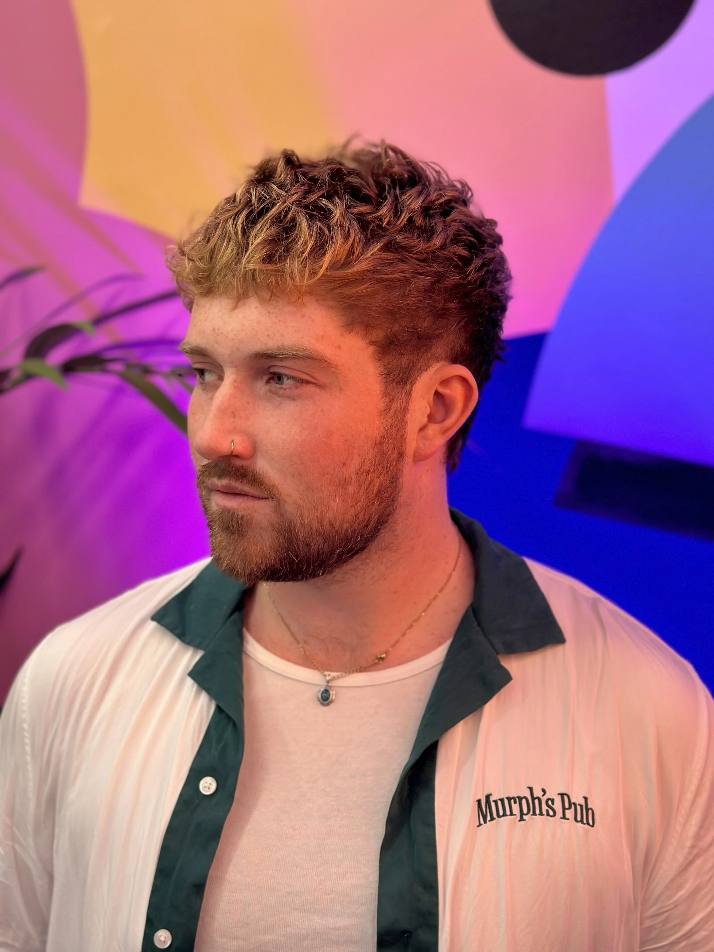 A young man with curly hair and a beard, wearing a white shirt with dark green accents, a necklace with a pendant, and a nose ring, standing against a colorful, abstract background with pink, purple, and blue hues.