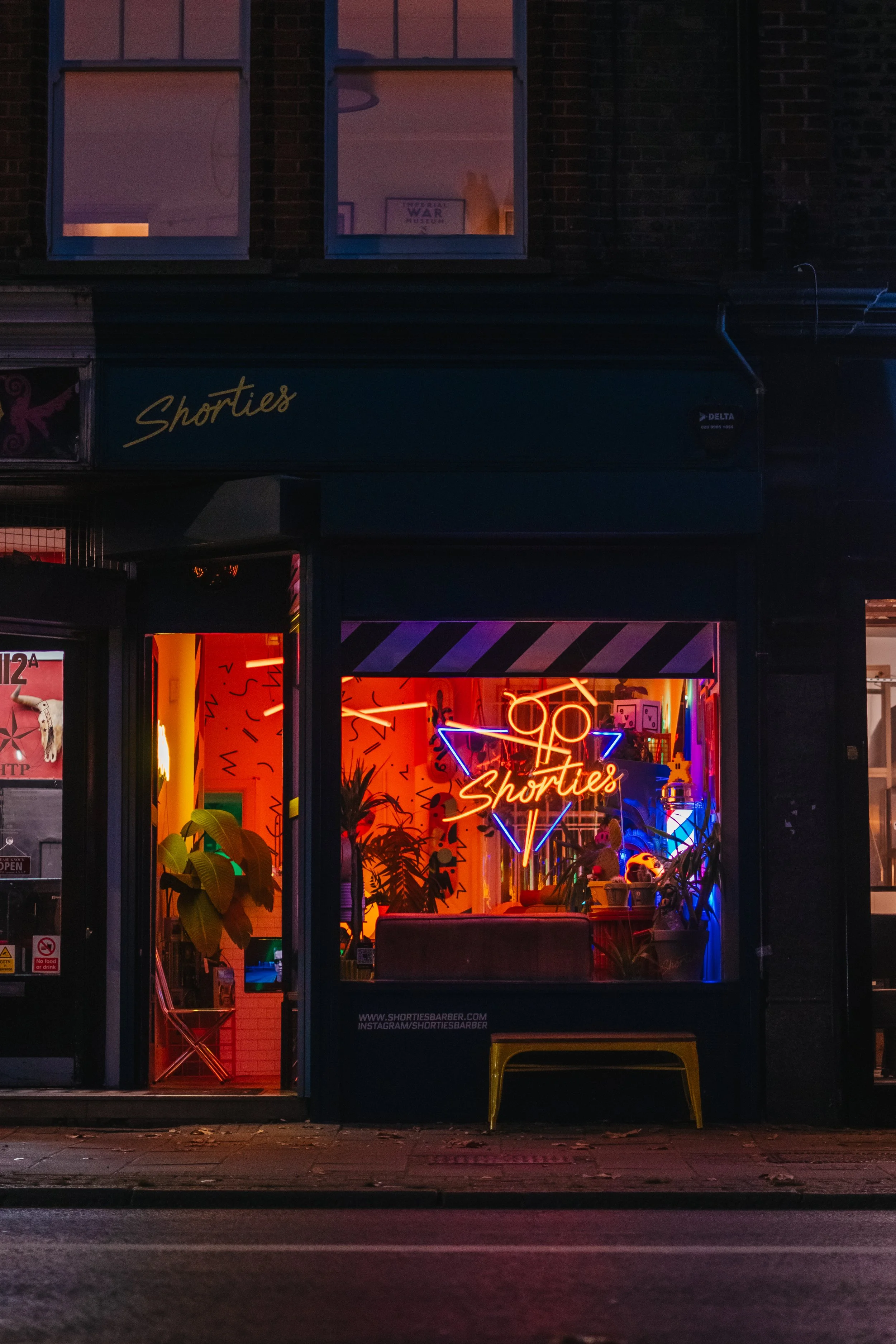A neon sign with a pair of scissors and the word 'Shorties' inside a store window, decorated with plants and colorful lights.