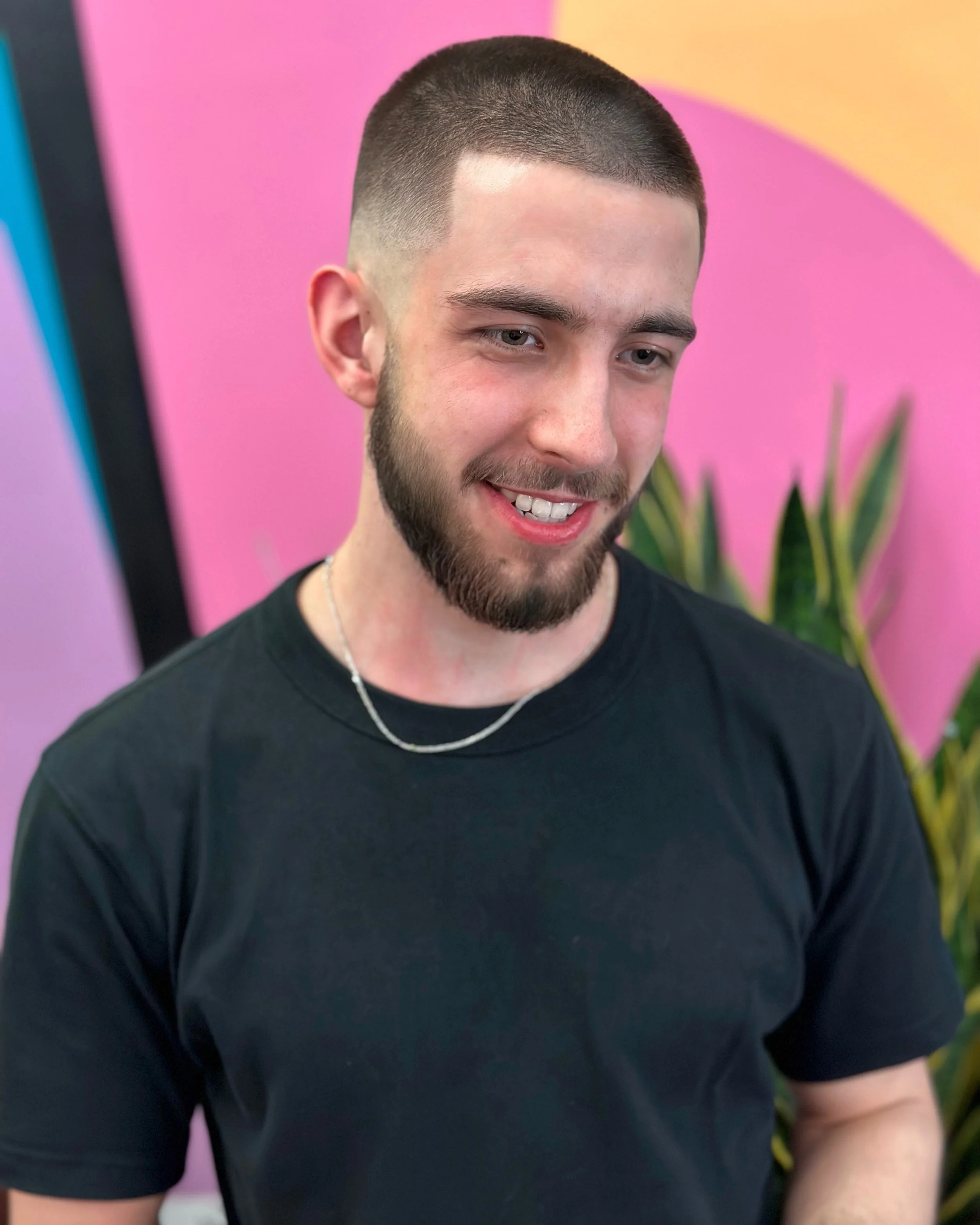 A young man with short, dark hair and a beard smiling in front of a colorful pink and purple background with a green plant visible behind him, wearing a black t-shirt and a thin silver necklace.