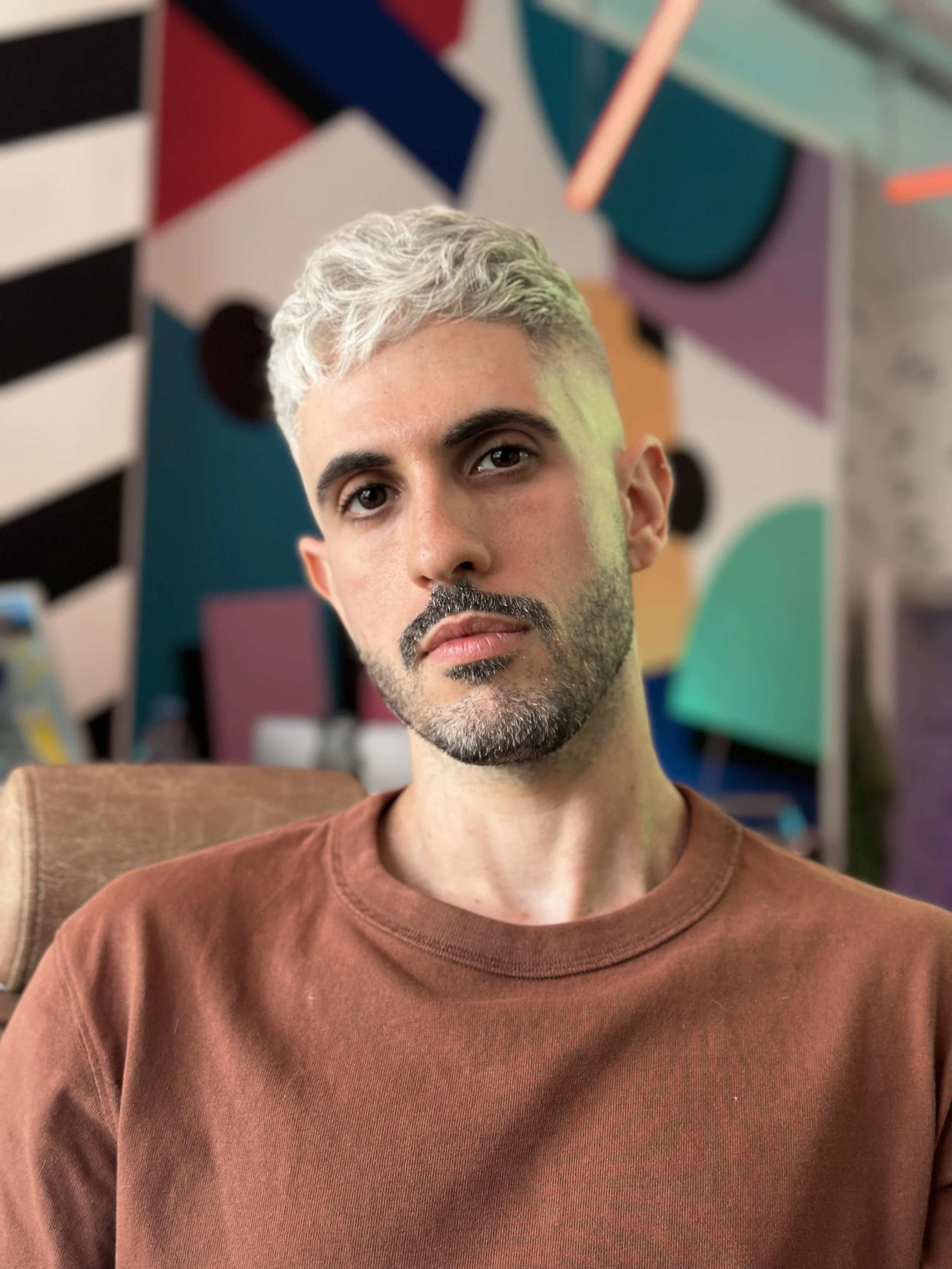 A man with short, black and platinum blonde hair, and a beard, wearing a brown shirt, sitting in front of a colorful abstract mural.