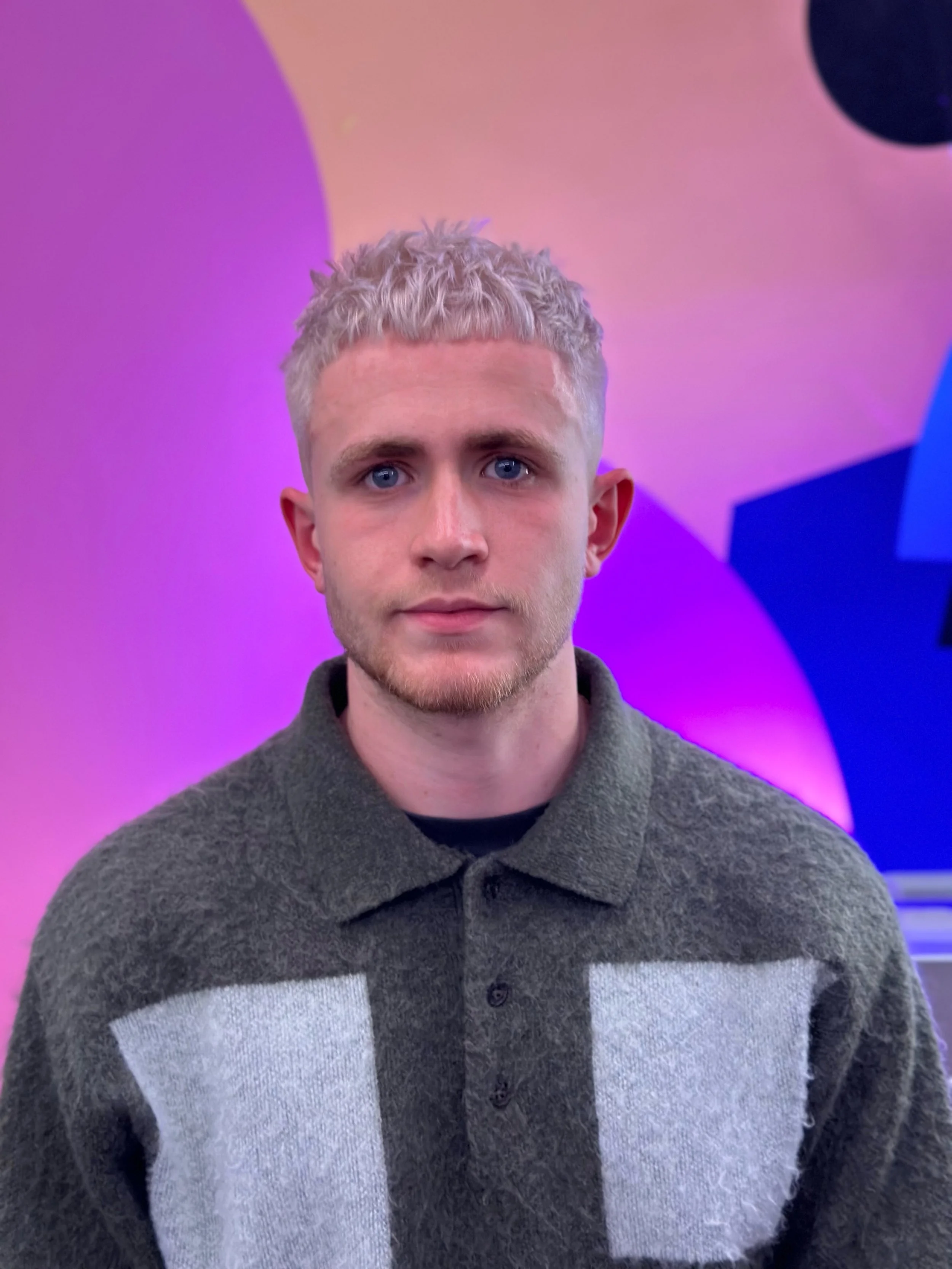 A young man with blond, curly hair and blue eyes standing in front of a colorful, geometric background.