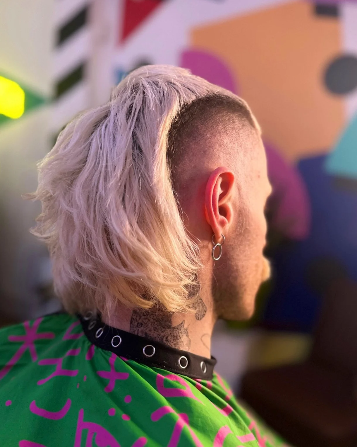 Side view of a person with short sides and long, platinum blonde, wavy hair on top, wearing a black choker and a green garment with purple writing, and an earring in the left ear; background features colorful abstract shapes.