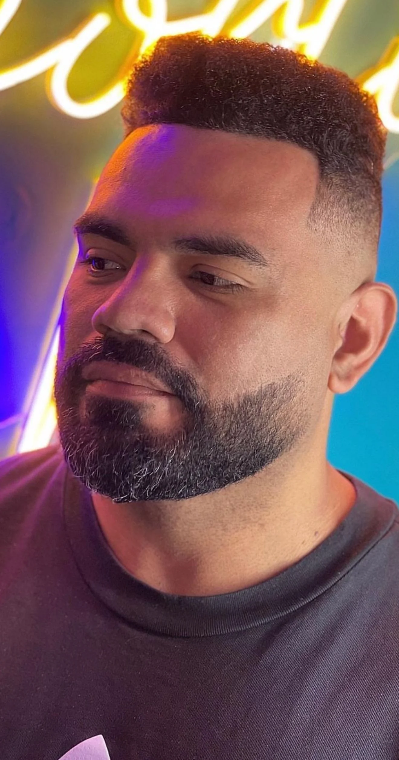 Close-up of a man with a beard and short hair, wearing a black shirt, looking slightly to the side with a neutral expression, against a colorful neon-lit background.