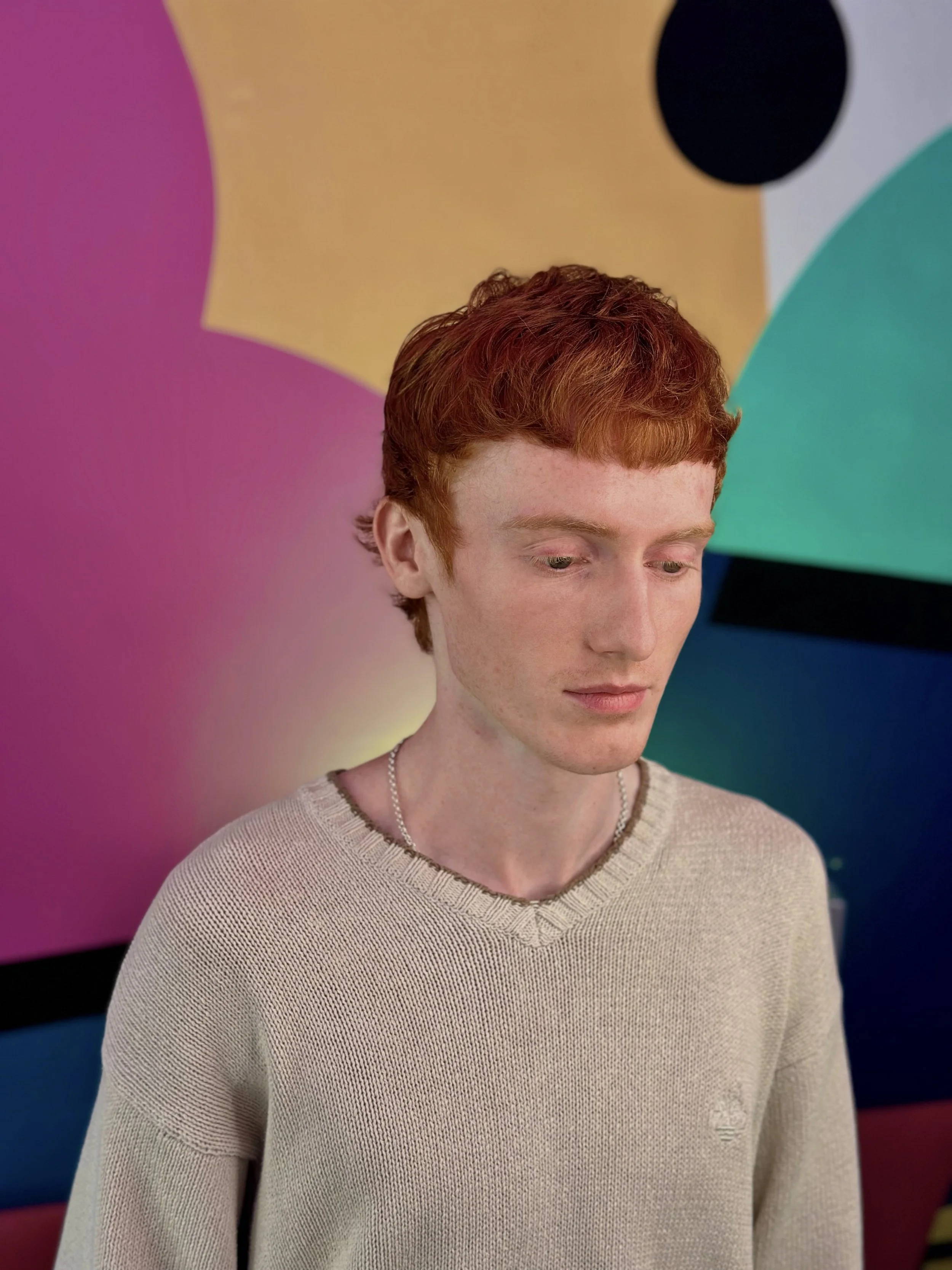A young man with red hair, wearing a beige sweater and a silver chain necklace, looking down, standing in front of a colorful abstract background with pink, yellow, blue, green, and black shapes.