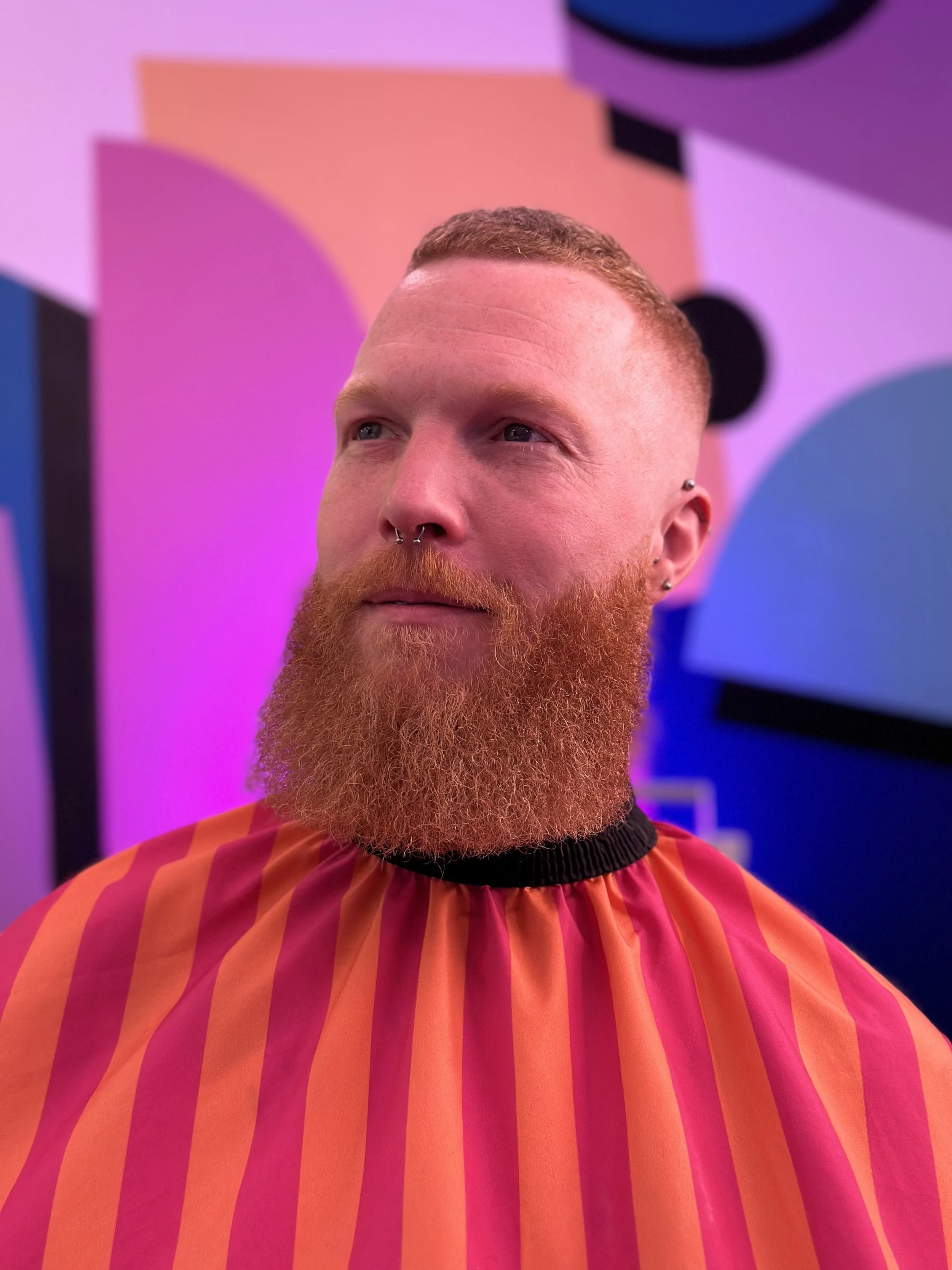 A man with a red beard and short hair, wearing a barber cape with red and pink stripes, in a colorful design background with pink, purple, and black abstract shapes.