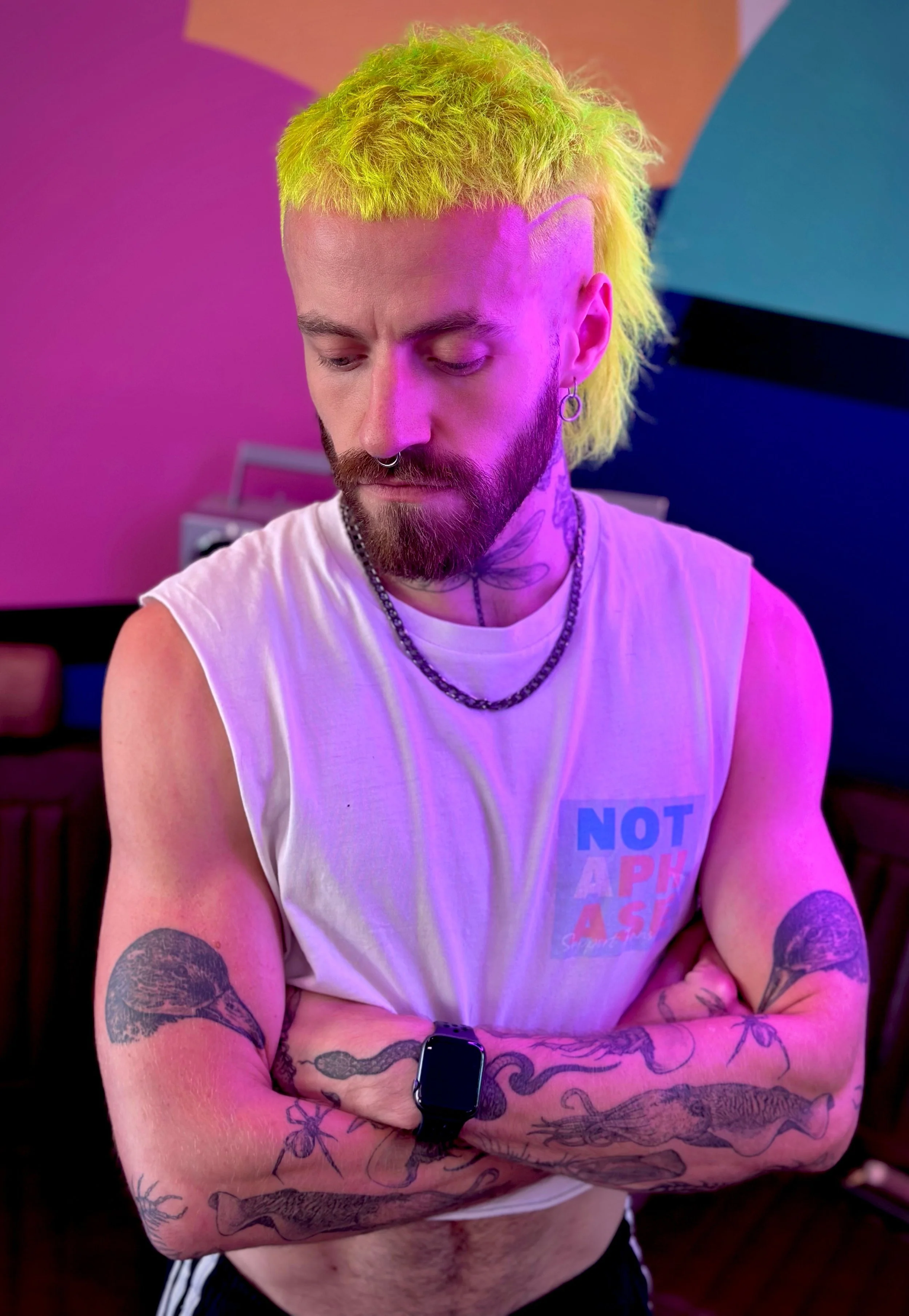 A young man with dyed yellow hair and tattoos on his arms, wearing a sleeveless shirt and a smartwatch, standing with arms crossed in a colorful room with pink, purple, and blue lighting.