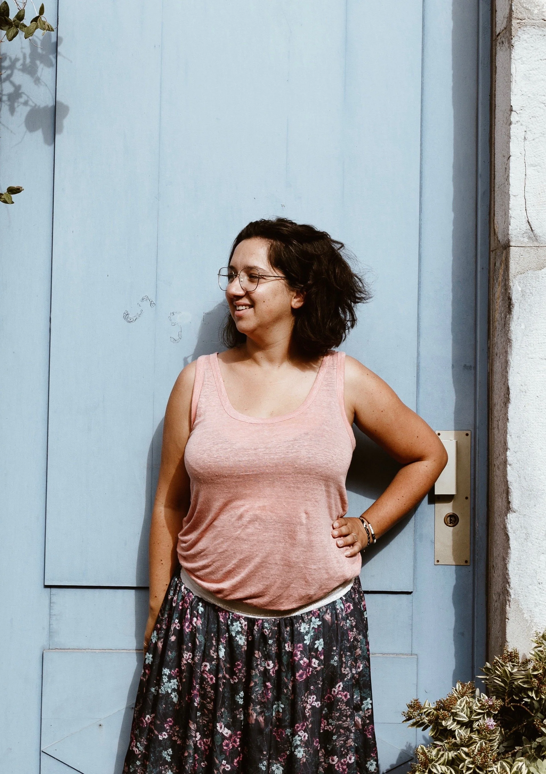 Jeune femme souriante avec lunettes, portant un débardeur rose et une jupe longue à motifs floraux, posant devant une porte bleue, avec des plantes en fleurs à droite.