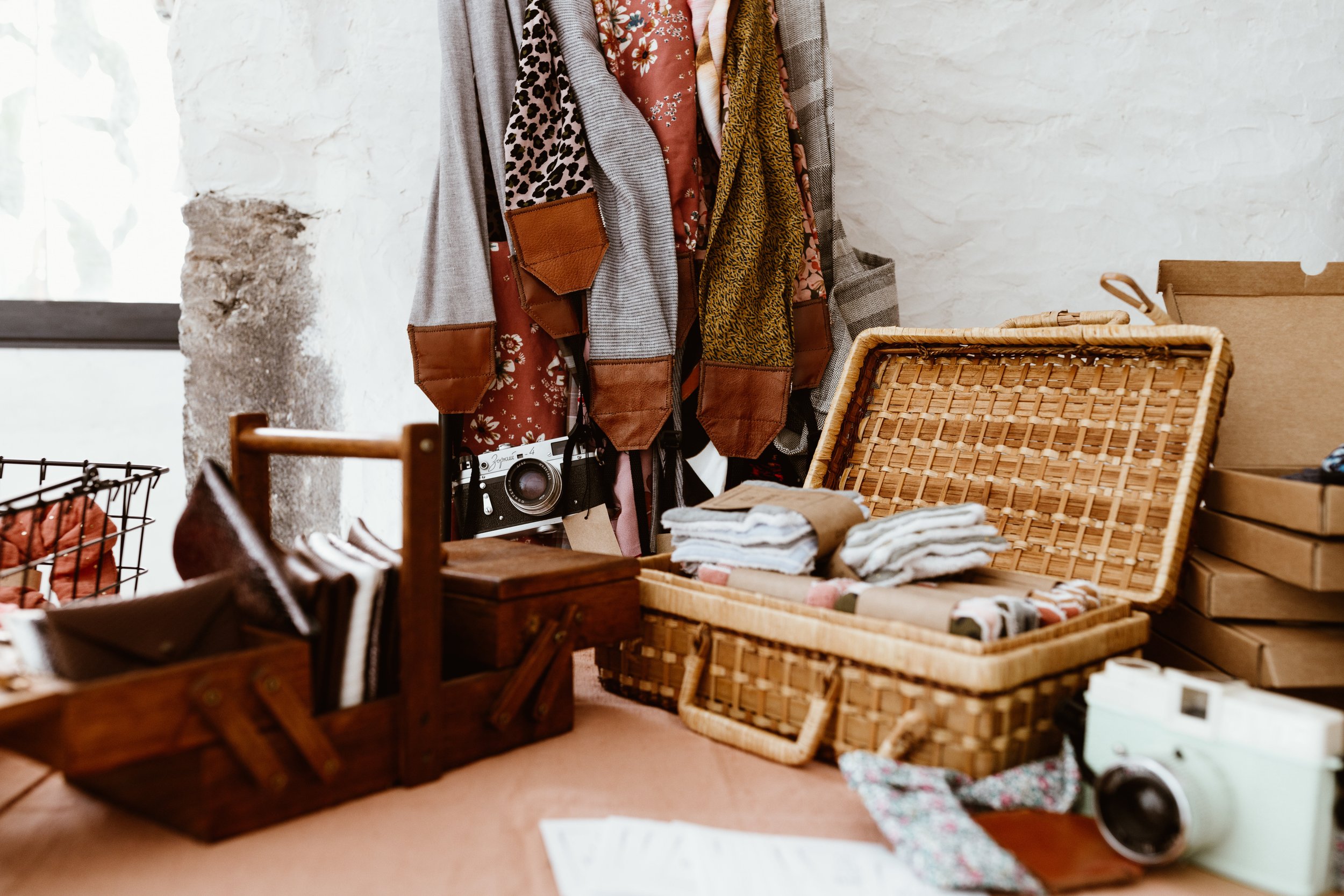 Une table avec des vêtements pliés, des boîtes en carton, un panier en osier, et d'autres objets dans un endroit ressemblant à une brocante ou un vide-grenier.