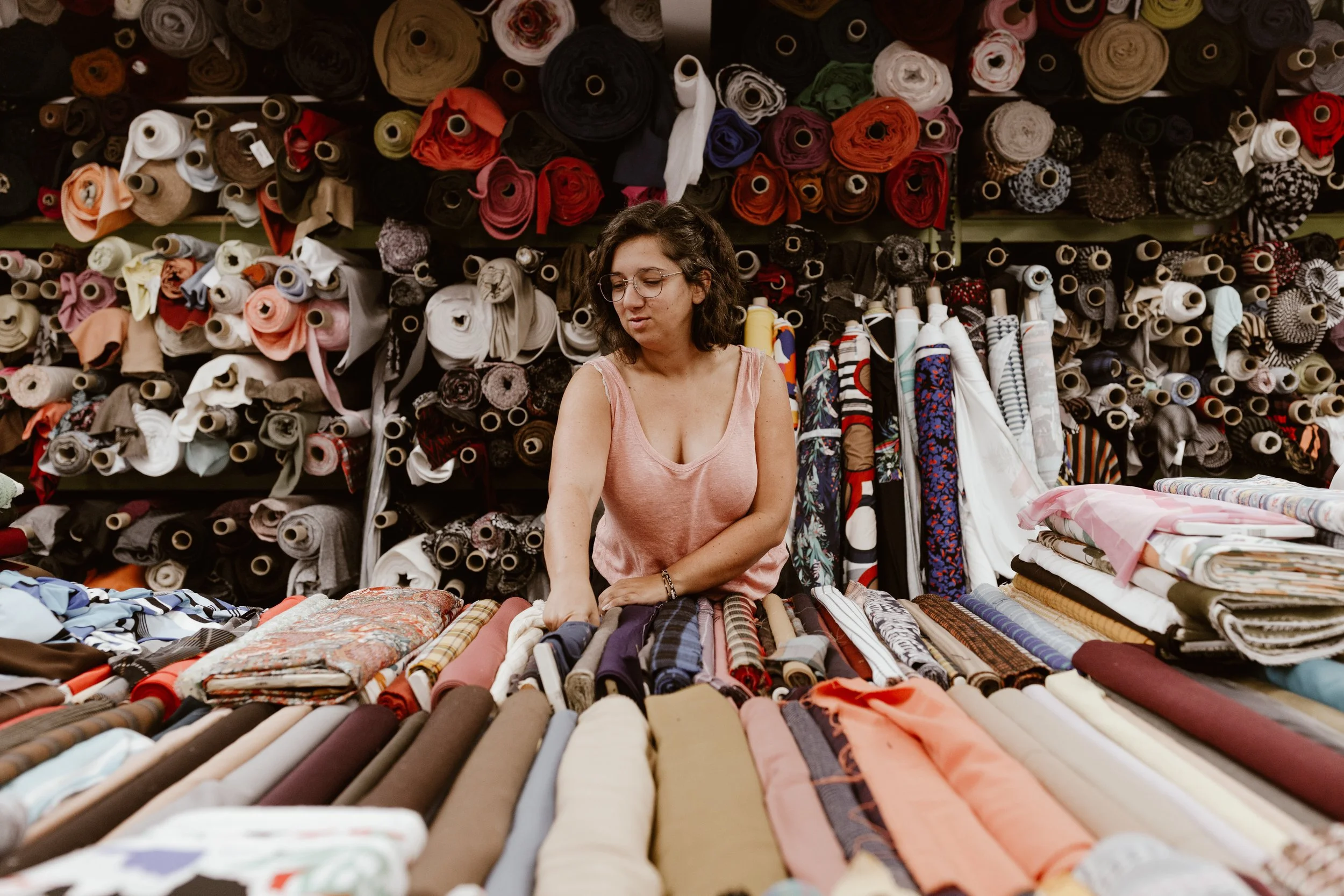 Une femme est en train de choisir des tissus dans un magasin de tissus, avec de nombreux rouleaux de tissus colorés derrière elle.