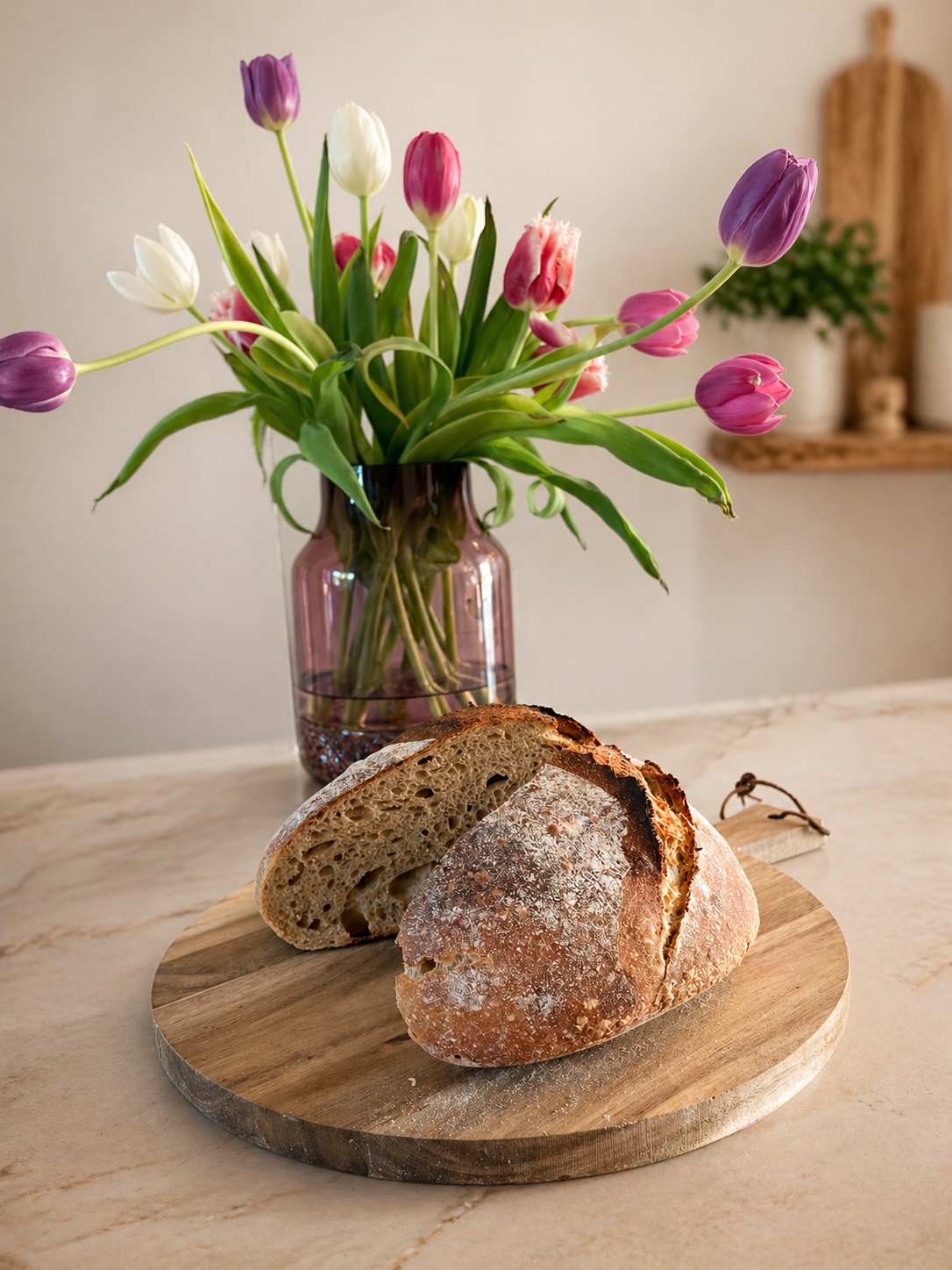Ein Laib Sauerteigbrot auf einem Holzbrett auf heller Küchenplatte mit Blumen im Hintergrund
