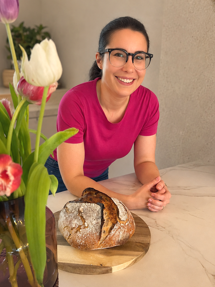 Eine Frau mit dunklen Haaren, Brille und pinkem T-Shirt, die lacht und vor einem Laib Sauerteigbrot auf einem Holzbrett steht, im Hintergrund Blumen in Vasen.