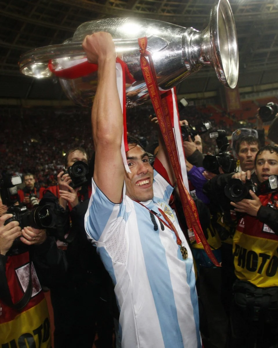 A man in a light blue and white soccer jersey holding a large silver trophy high above his head, smiling, surrounded by photographers in a stadium.
