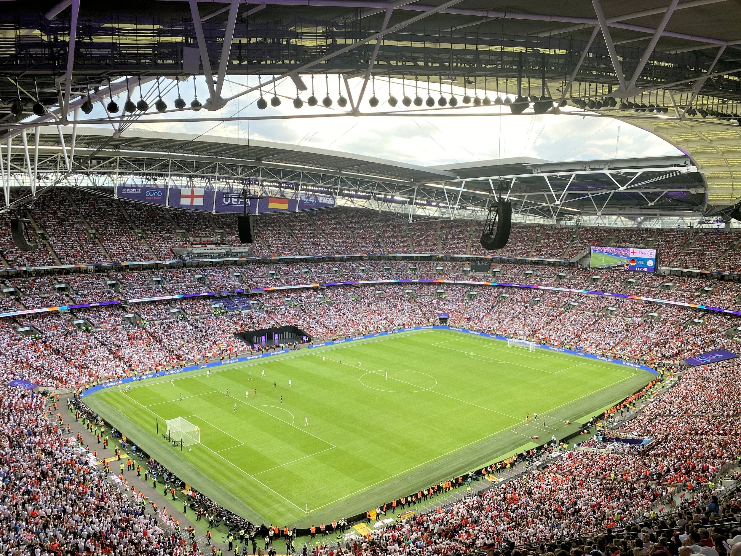 A large football stadium filled with spectators watching a match, with players on the field and banners hanging from the ceiling.