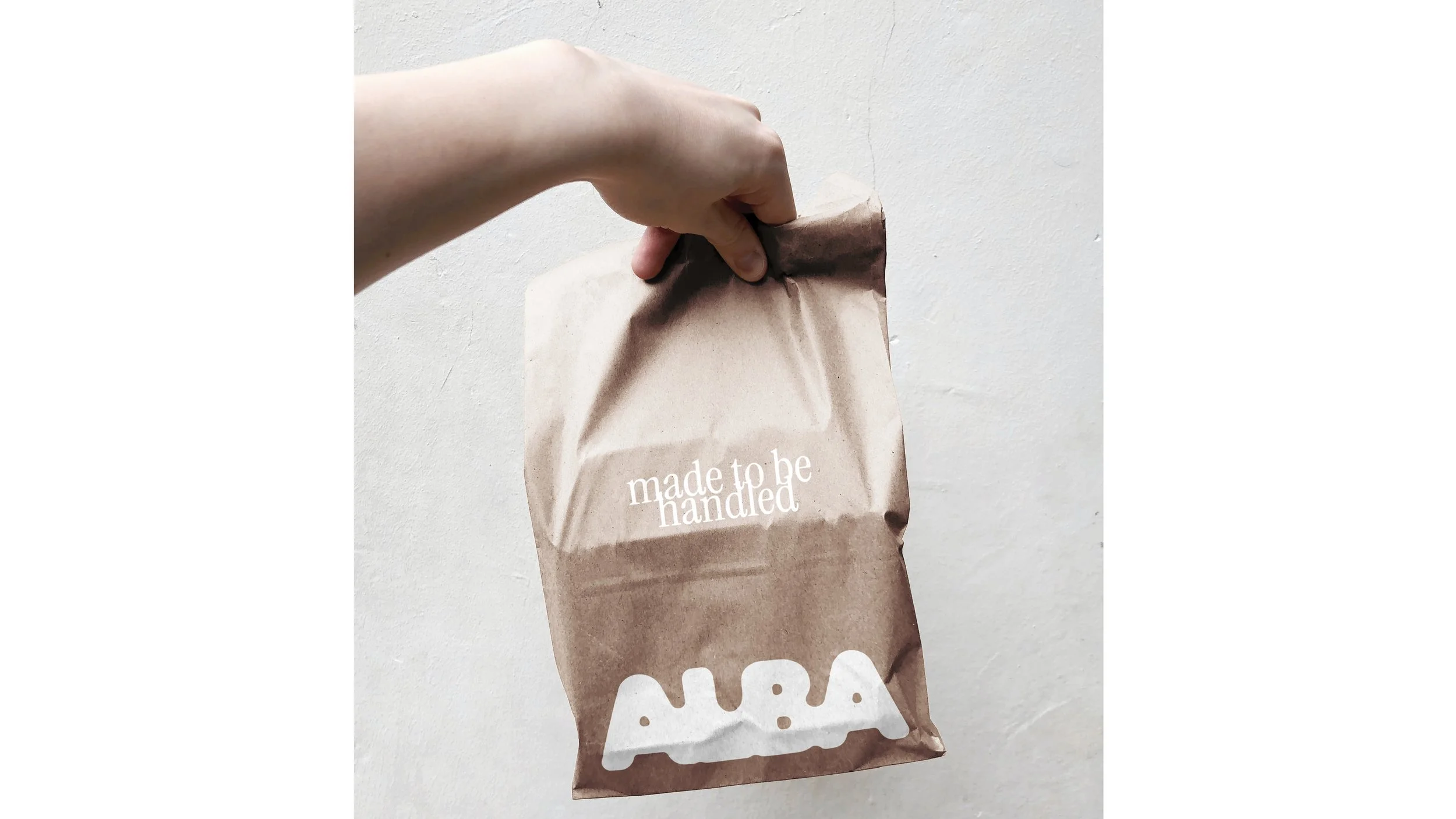Brown paper bag with the words "made to be handled" printed on it, being held by a hand against a plain light-colored wall.
