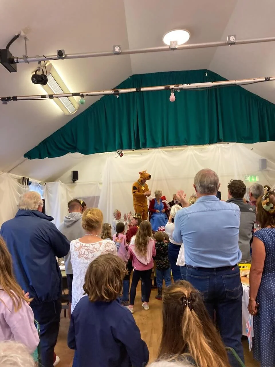 Children and adults watching a performance on a stage with a person in a horse costume and another in a blue dress, inside a decorated event hall with a green curtain backdrop.